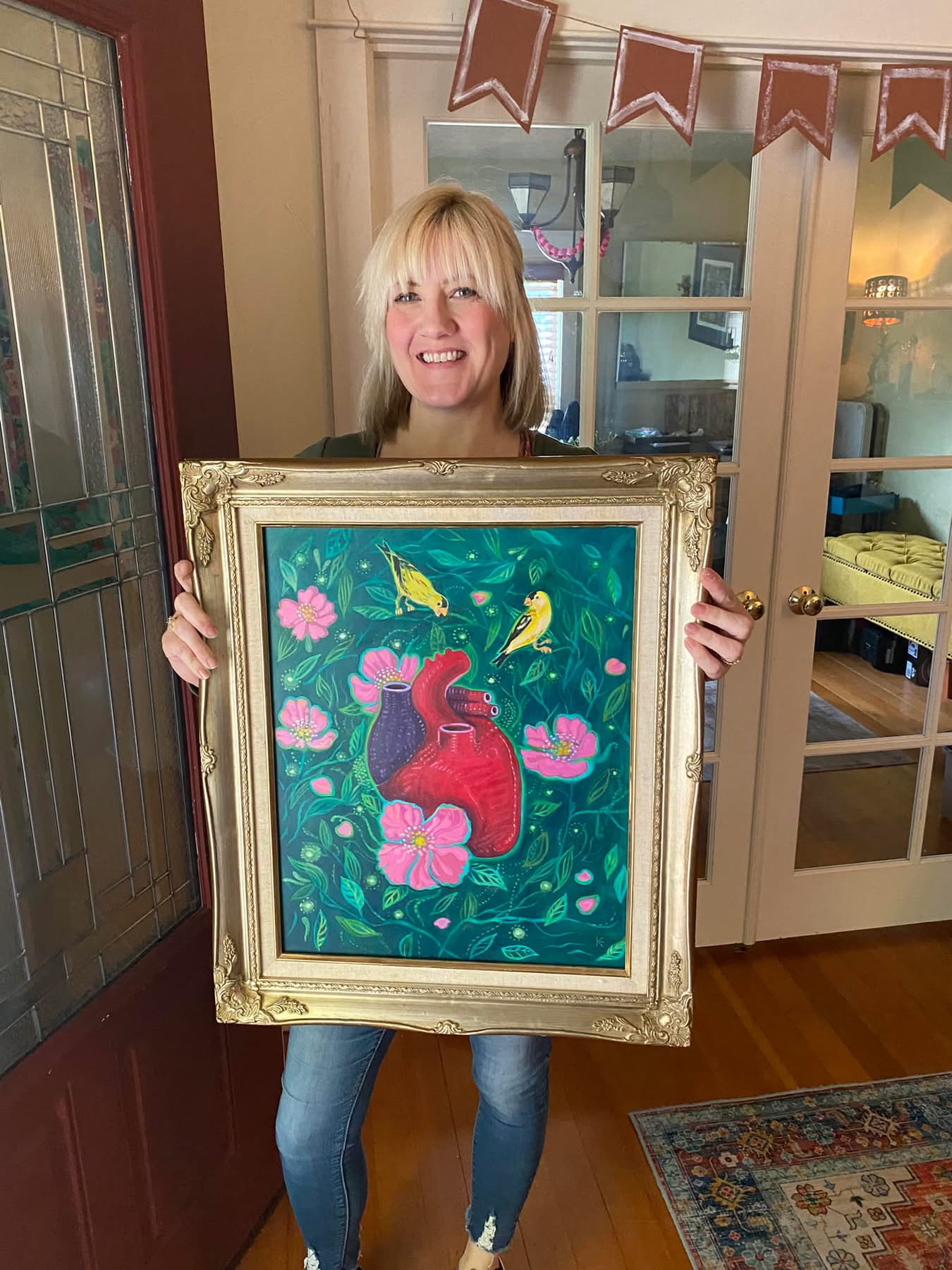 Woman with blonde hair smiling, holding a framed colorful painting of a red teapot surrounded by pink flowers and yellow butterflies.