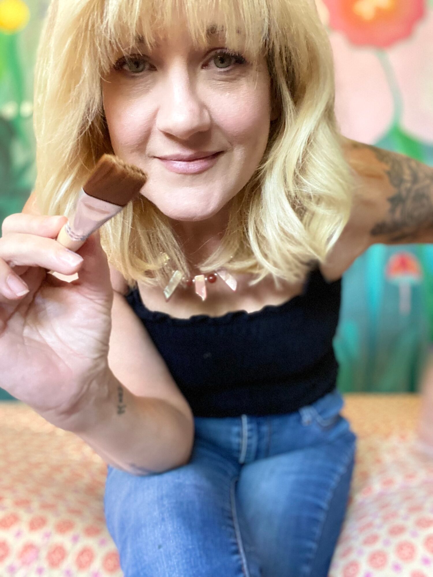 Woman with blonde hair holding a paintbrush, smiling, in a colorful room with floral decorations.