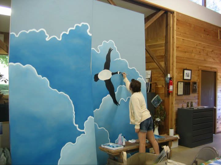 Person painting a large mural of a whale in the sky with clouds on a blue background, indoors.