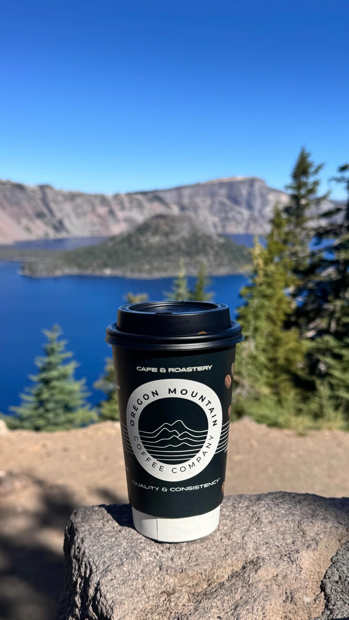 Coffee cup on rock with lake, trees, and mountains in background under clear blue sky