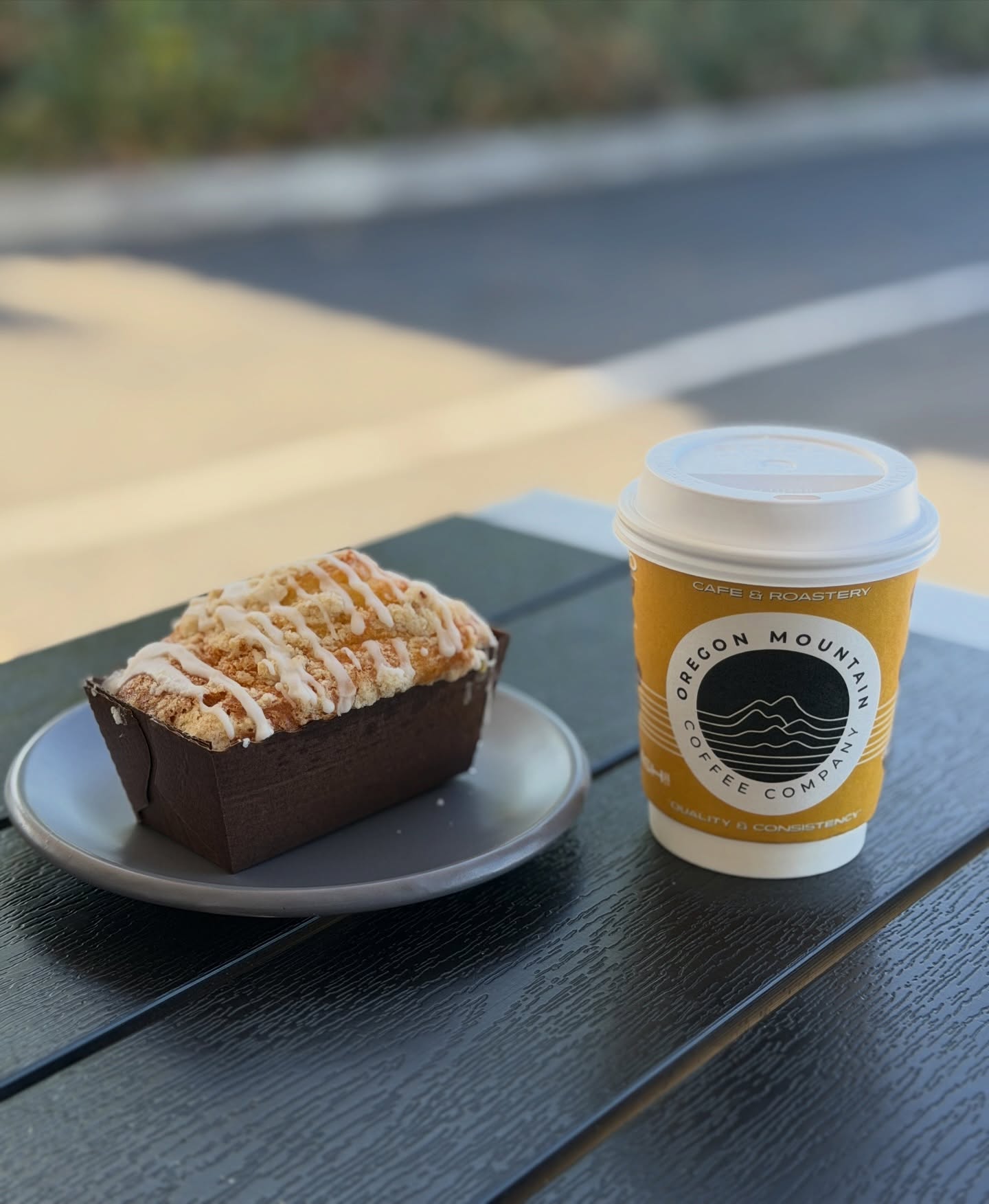 Coffee cup with Oregon Mountains logo and a pastry on a small plate on a dark wooden table outdoors.