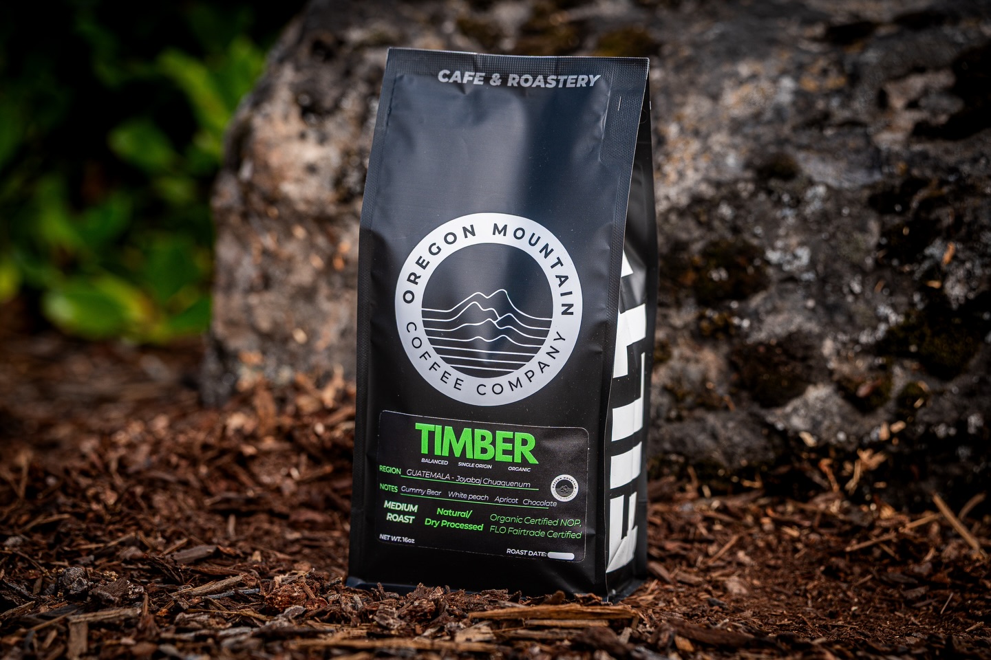 Black bag of coffee with white and green text, placed on soil near a rock, outdoors, with greenery in background.
