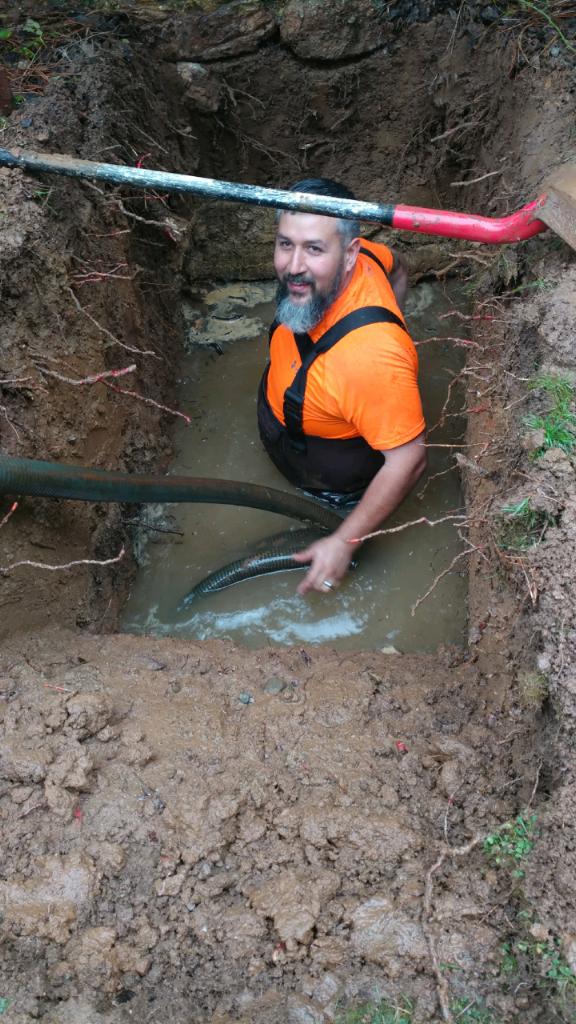 Man with beard in orange shirt and black vest stands in muddy water holding a black hose, with a shovel resting on his shoulder.