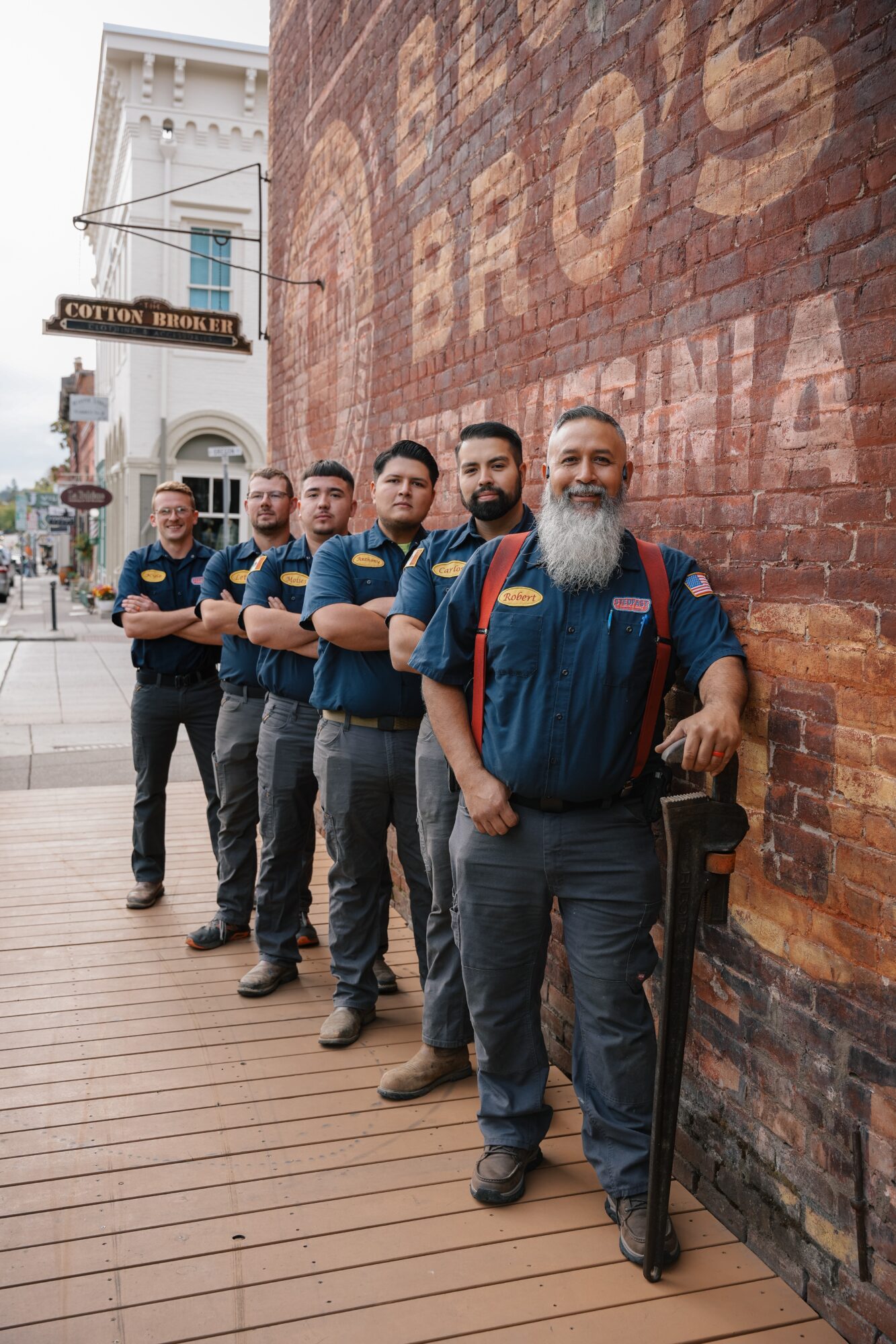 Six men stand in a line outside a brick building, some with arms crossed, smiling, with a sign reading 'Cotton Board' nearby.