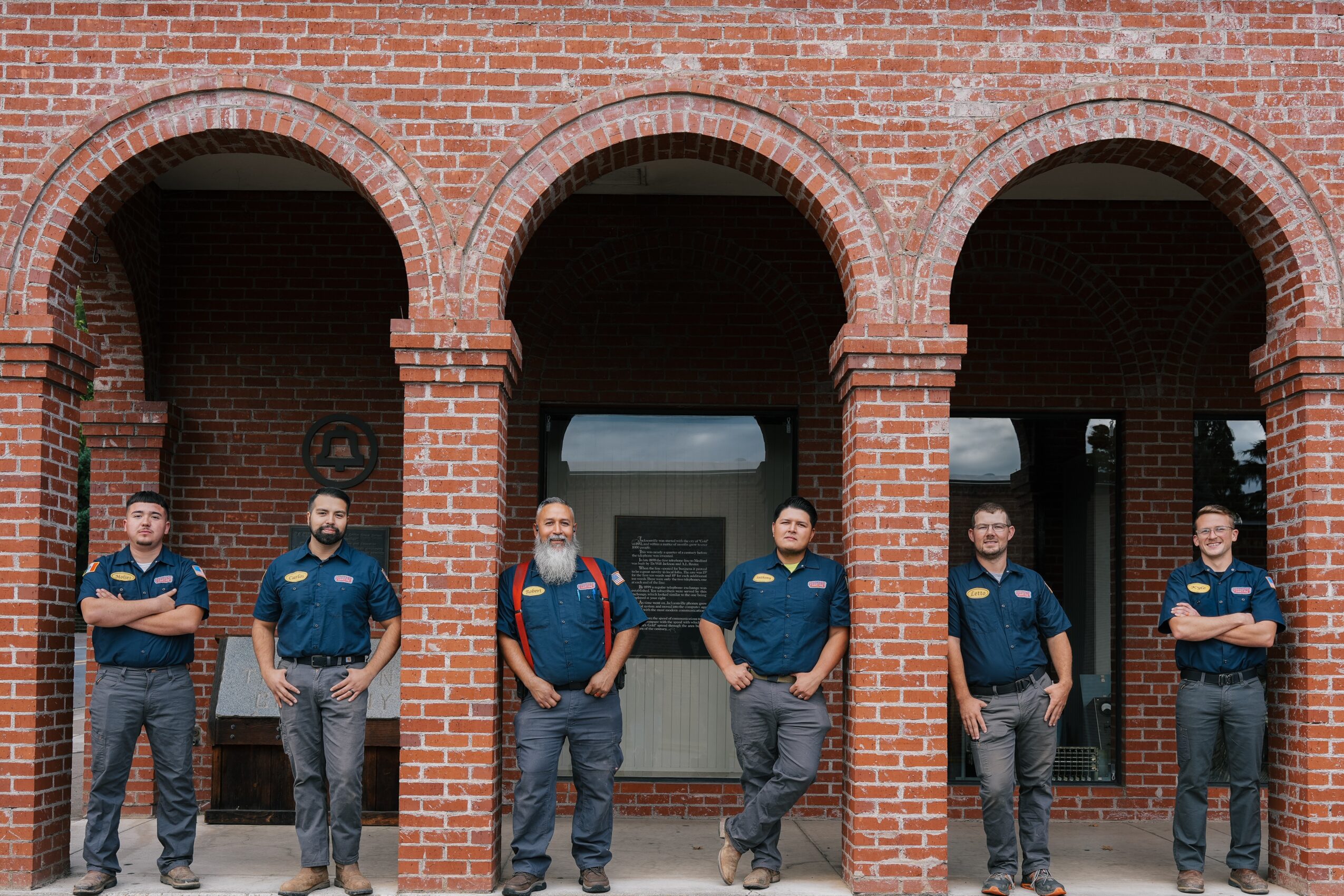 Six men stand in front of a brick building with arches, wearing dark blue shirts and jeans.