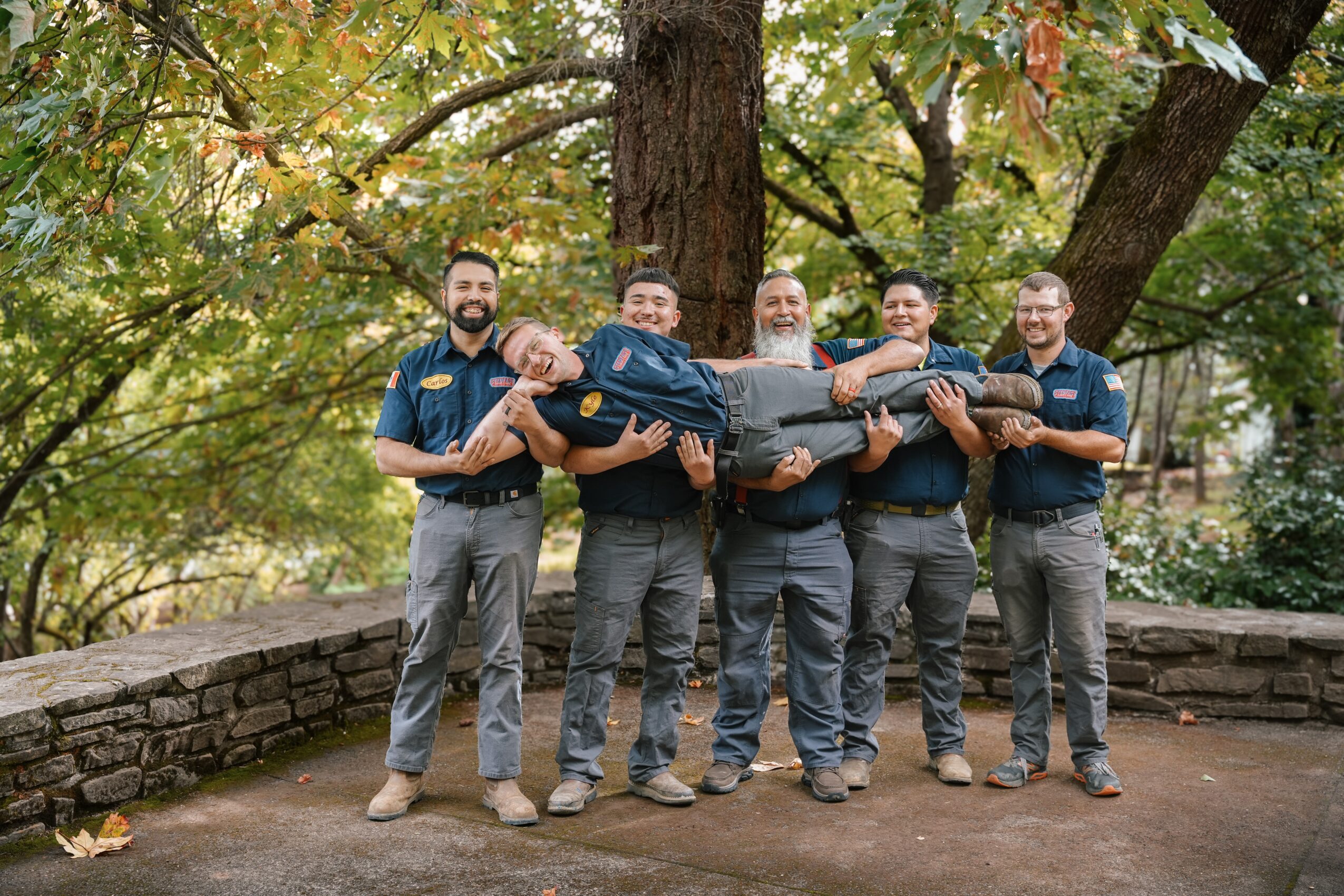 Five men outdoors holding a person horizontally, with trees and a stone wall in the background.