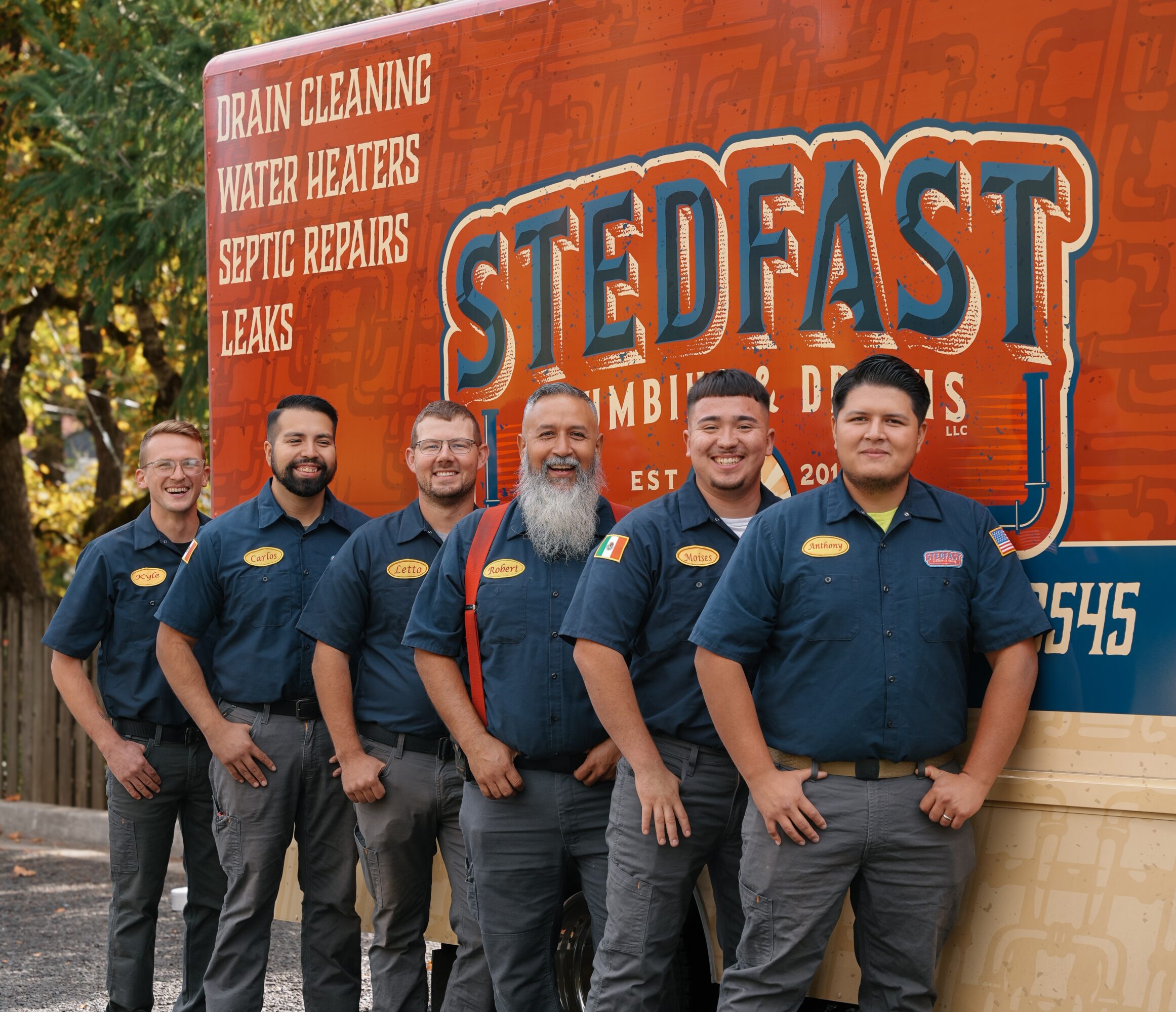 Five people standing in front of a Stedfast truck, smiling, wearing matching uniforms with name tags, outdoors with trees in background.
