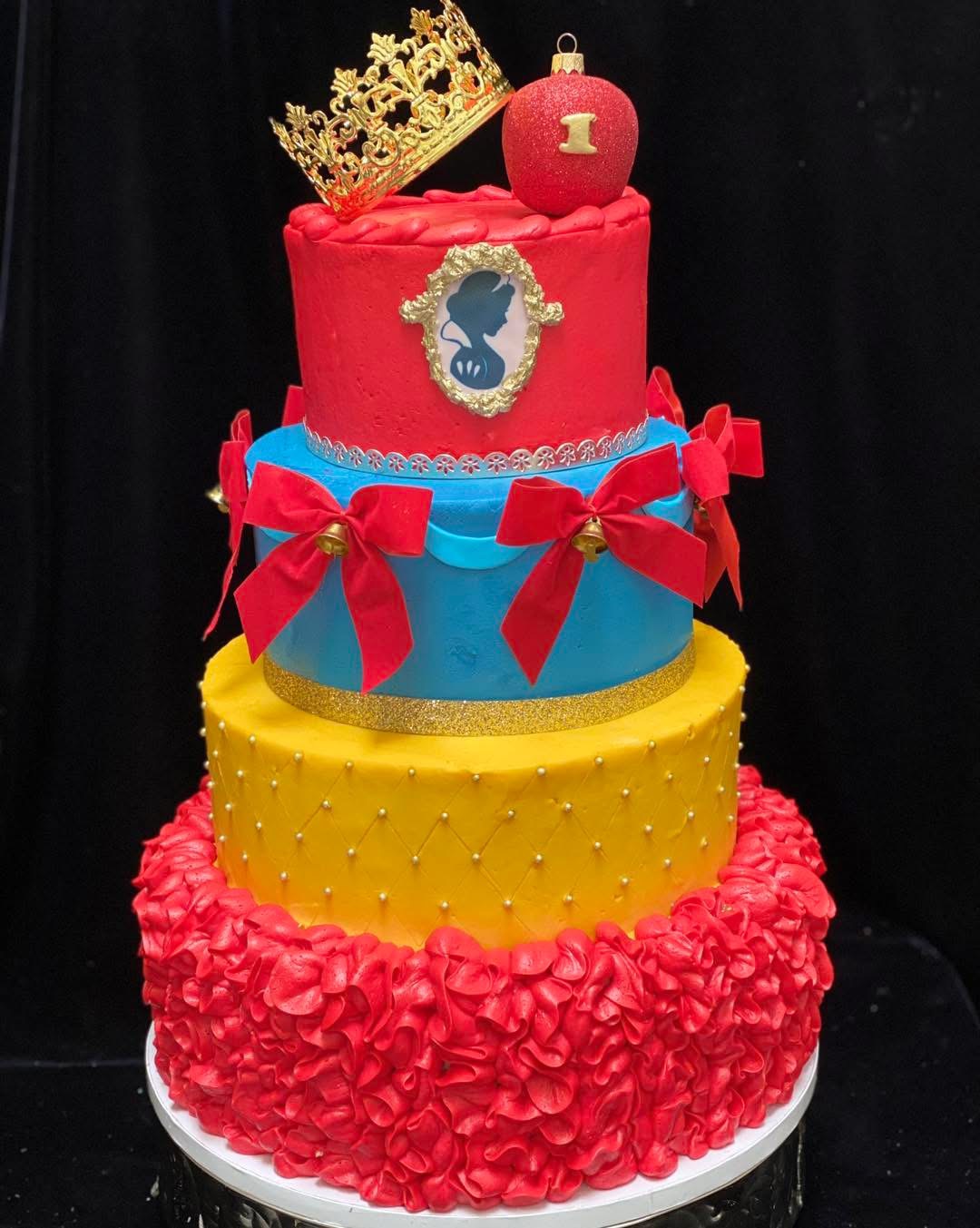 Four-tiered cake with a gold crown, pink, blue, yellow, and red decorations, and a pink ball on top.