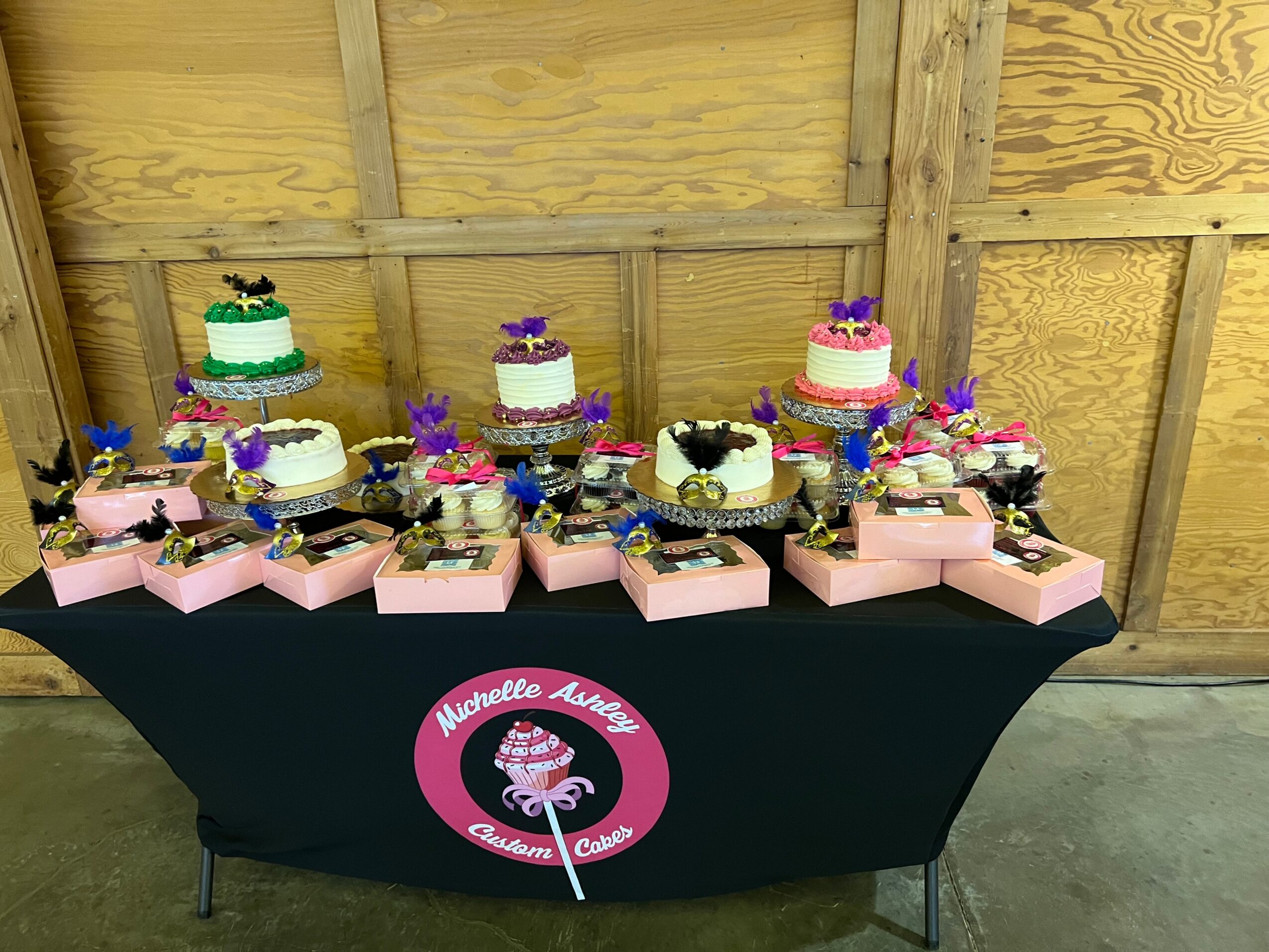 Table with decorated cupcakes and pink boxes, wooden wall background, pink circular sign with a cupcake logo, 20 words.