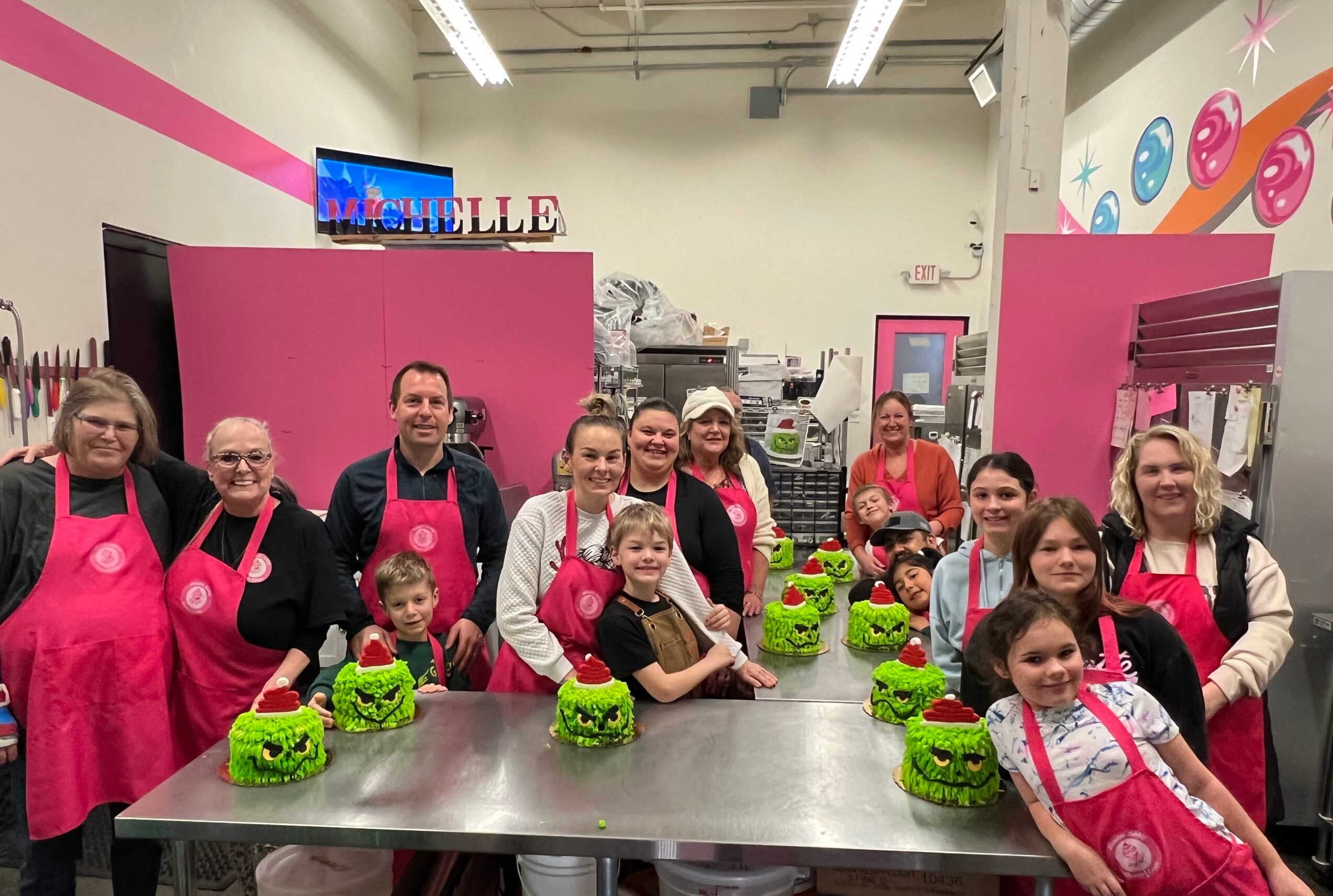 Group of children and adults in pink aprons around a table with green decorated cupcakes in a bakery.
