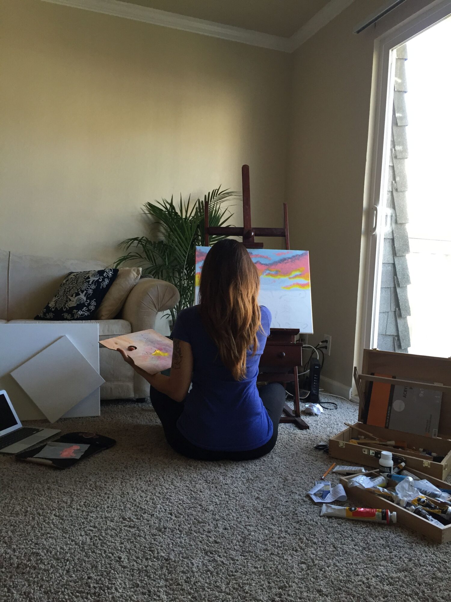 Person sitting on carpeted floor, painting on canvas, with art supplies and a window in background.