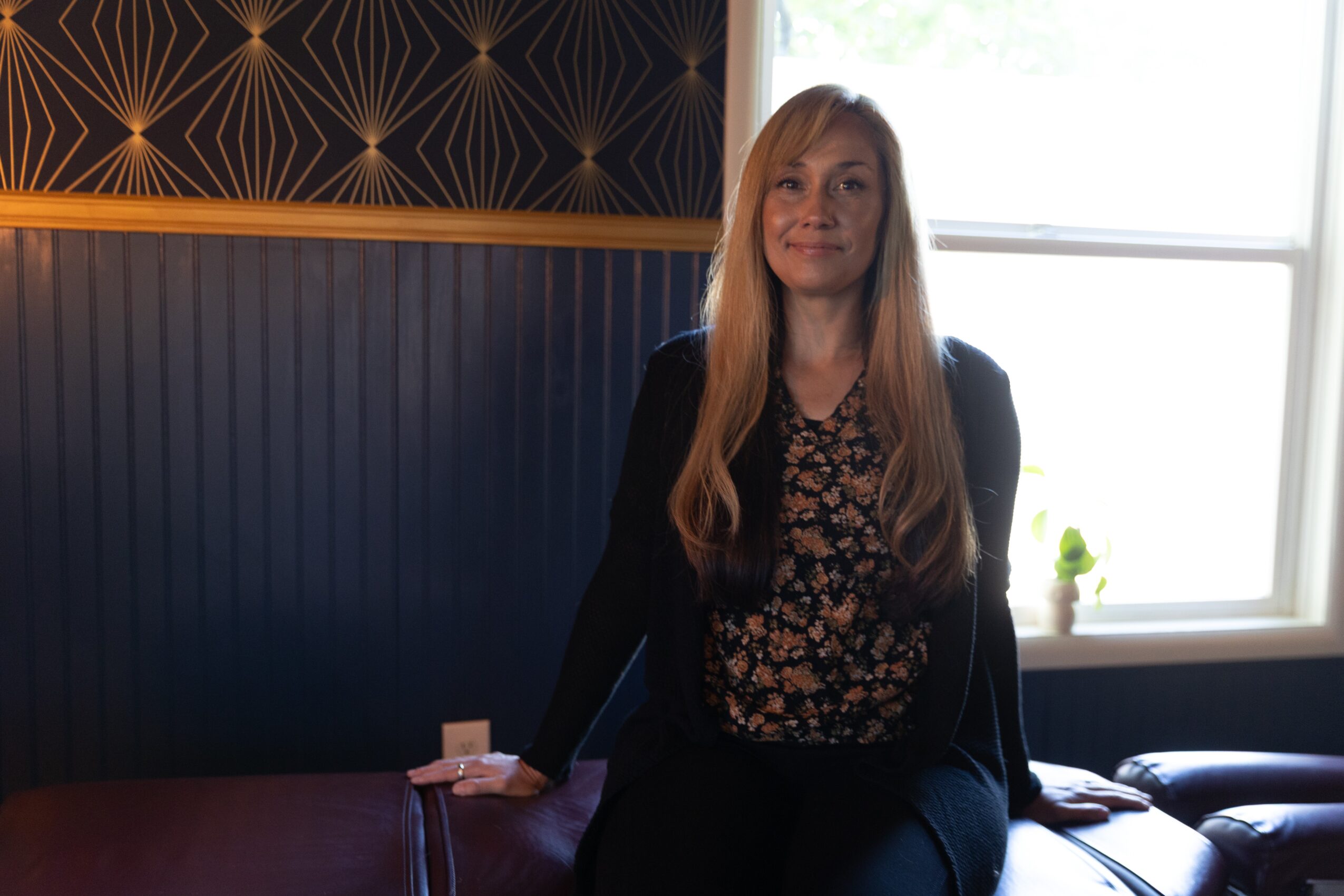 Woman with long blonde hair sitting by a window, wearing a black blazer and floral blouse, in a room with dark wall decor.