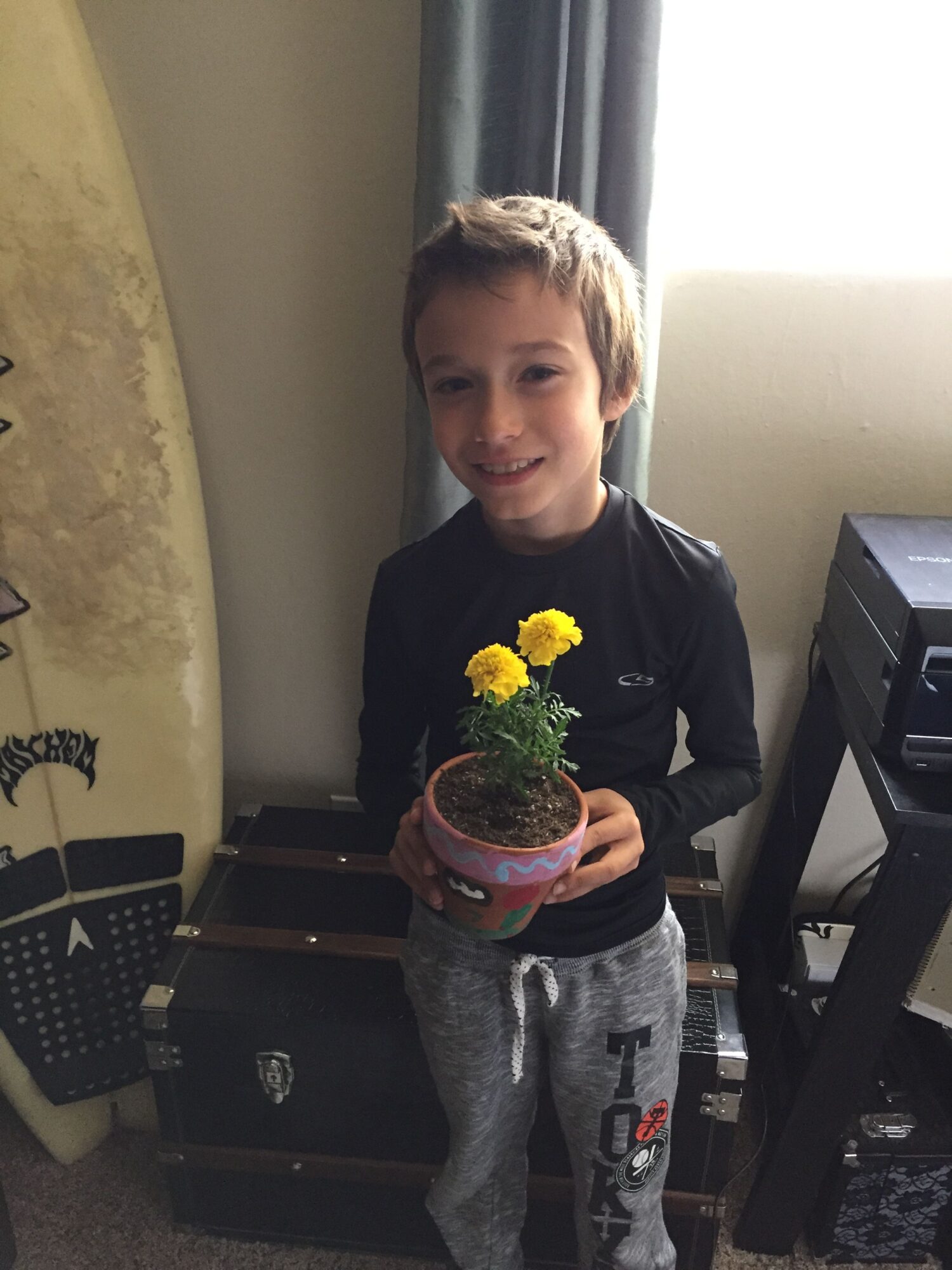 Boy holding a small potted plant with yellow flowers indoors, standing near a window with curtains.
