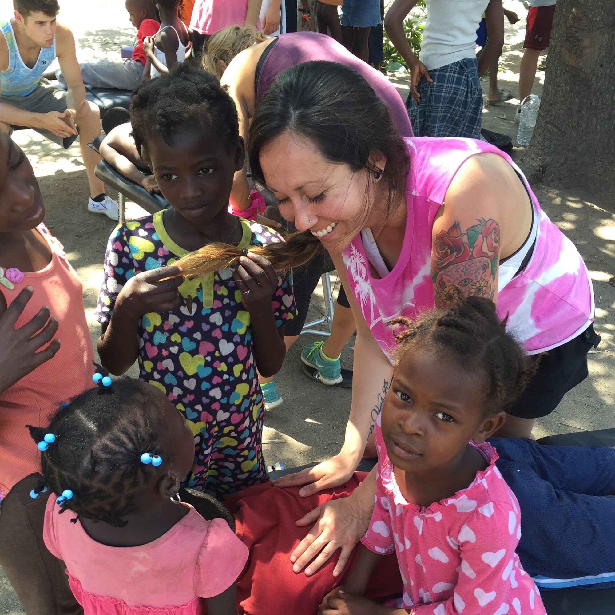 Smiling woman with tattoos interacts with children outdoors, some holding food, in a sunny setting with trees and other people.