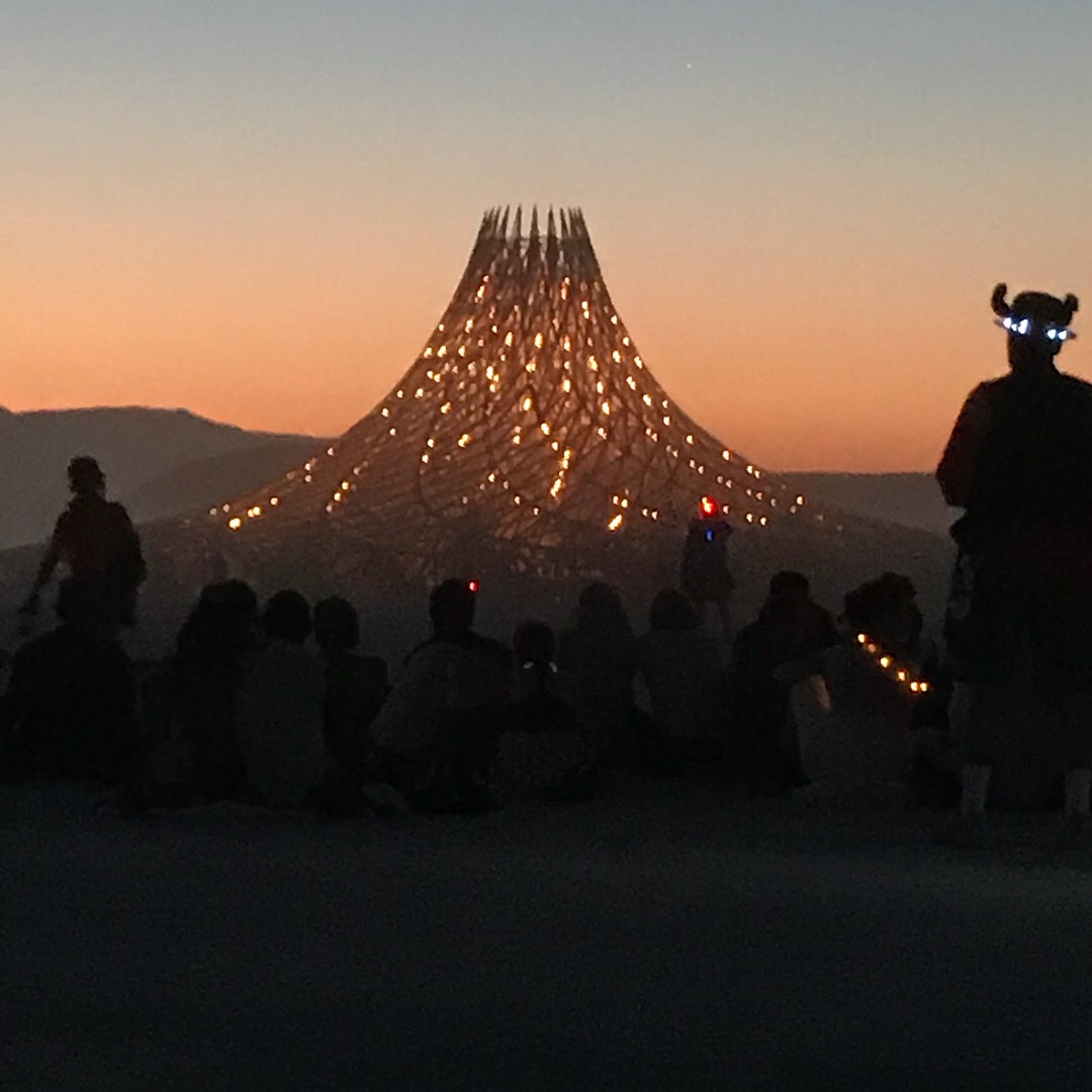 People sitting and standing outdoors at sunset with a large illuminated structure in the background.