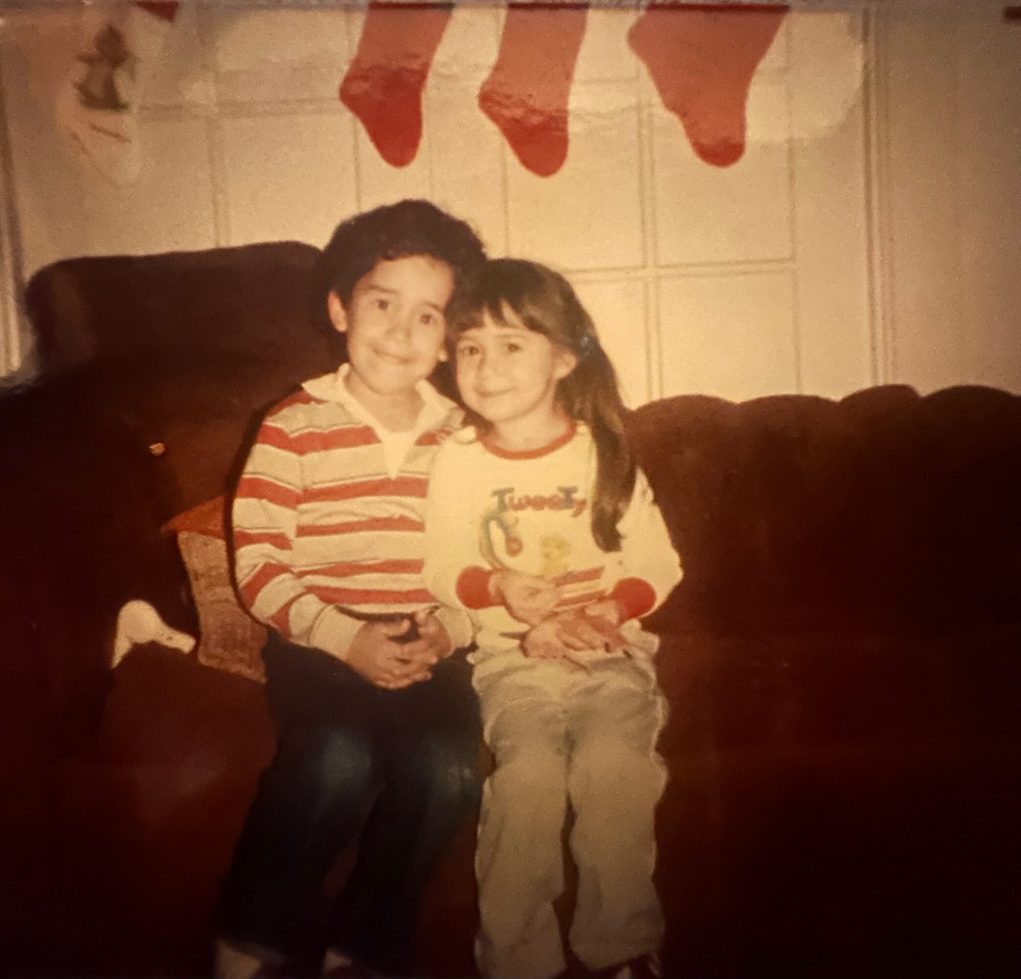 Two children sitting on a dark sofa, with Christmas stockings hanging above them, smiling at the camera.