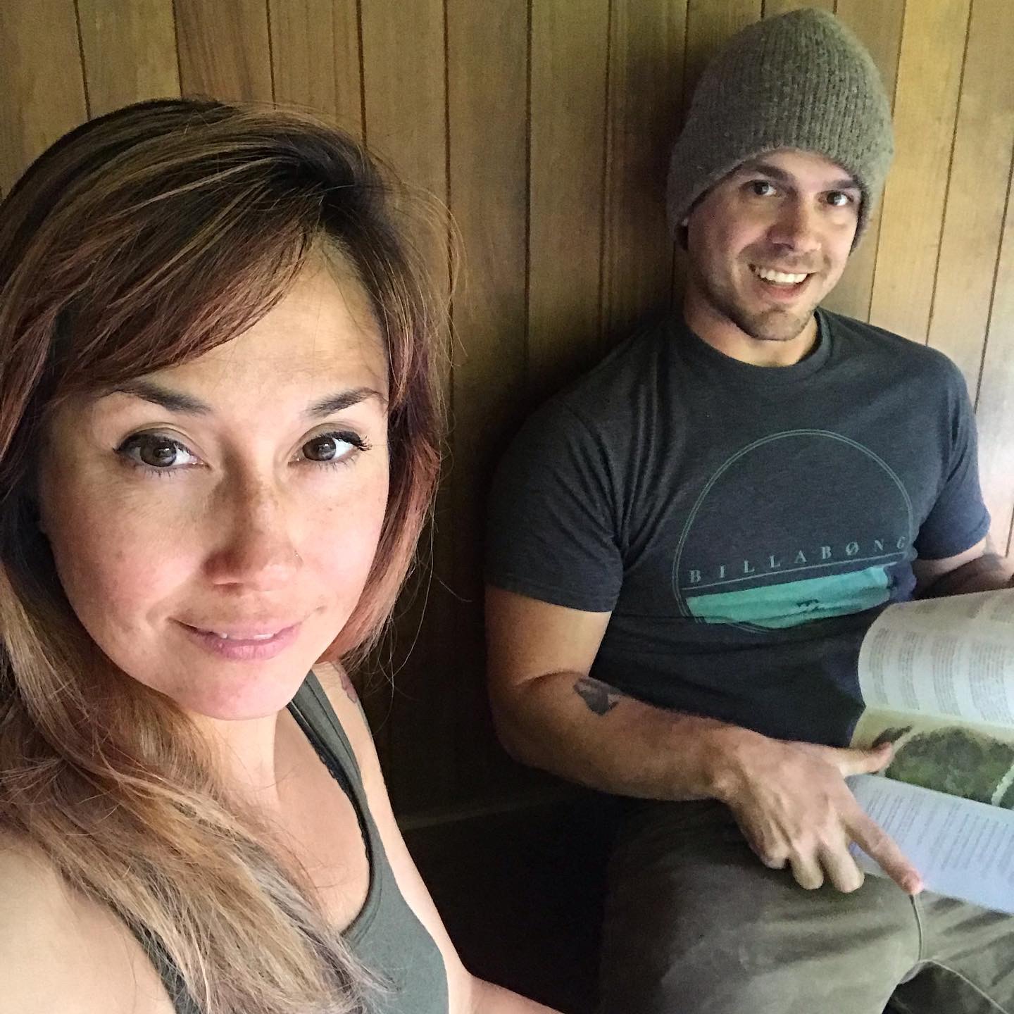 A woman and a man sitting against a wooden wall, woman smiling at the camera, man reading a book, wearing a beanie.
