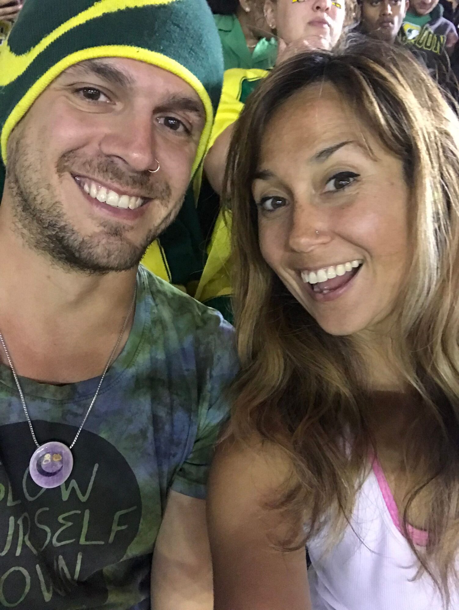 Two smiling people, a woman with long hair and a man wearing a hat, close together outdoors.