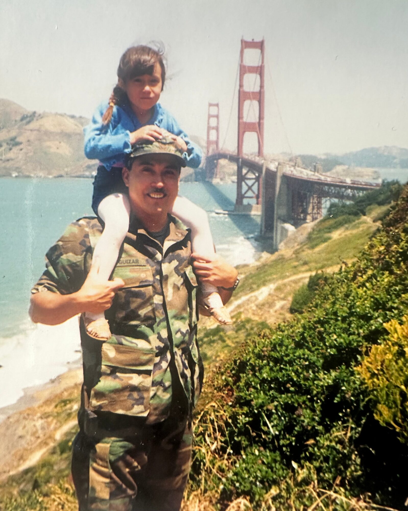 Child sitting on adult's shoulders near a bridge over water, with greenery in foreground and hills in background.