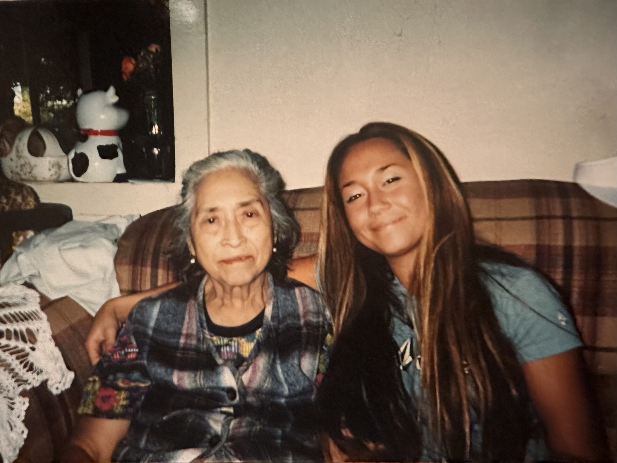 An elderly woman and a young girl sit together on a couch, smiling, with a window and decorations behind them.