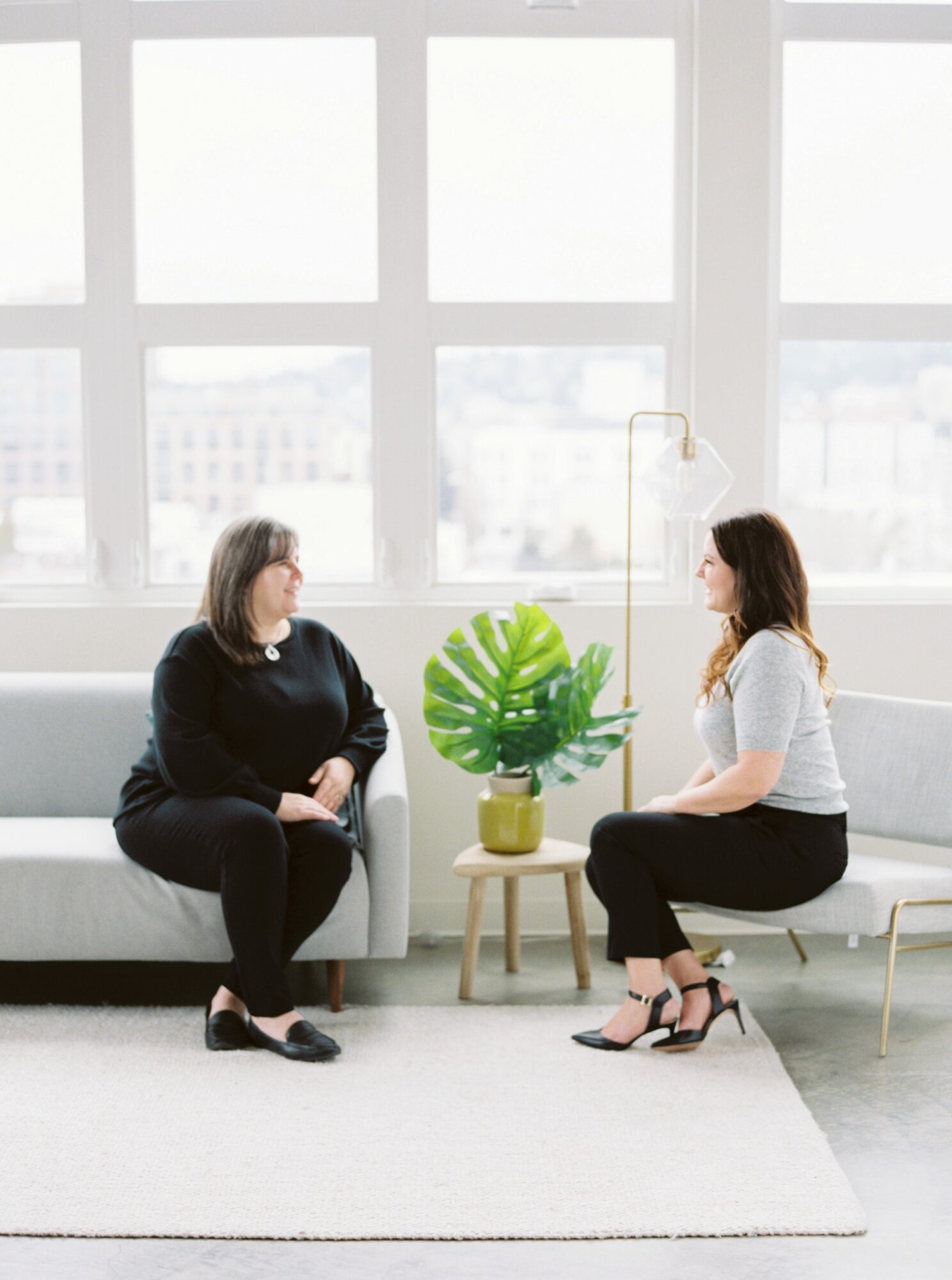 Two women sit facing each other in a bright room with large windows, a plant on a small table between them.