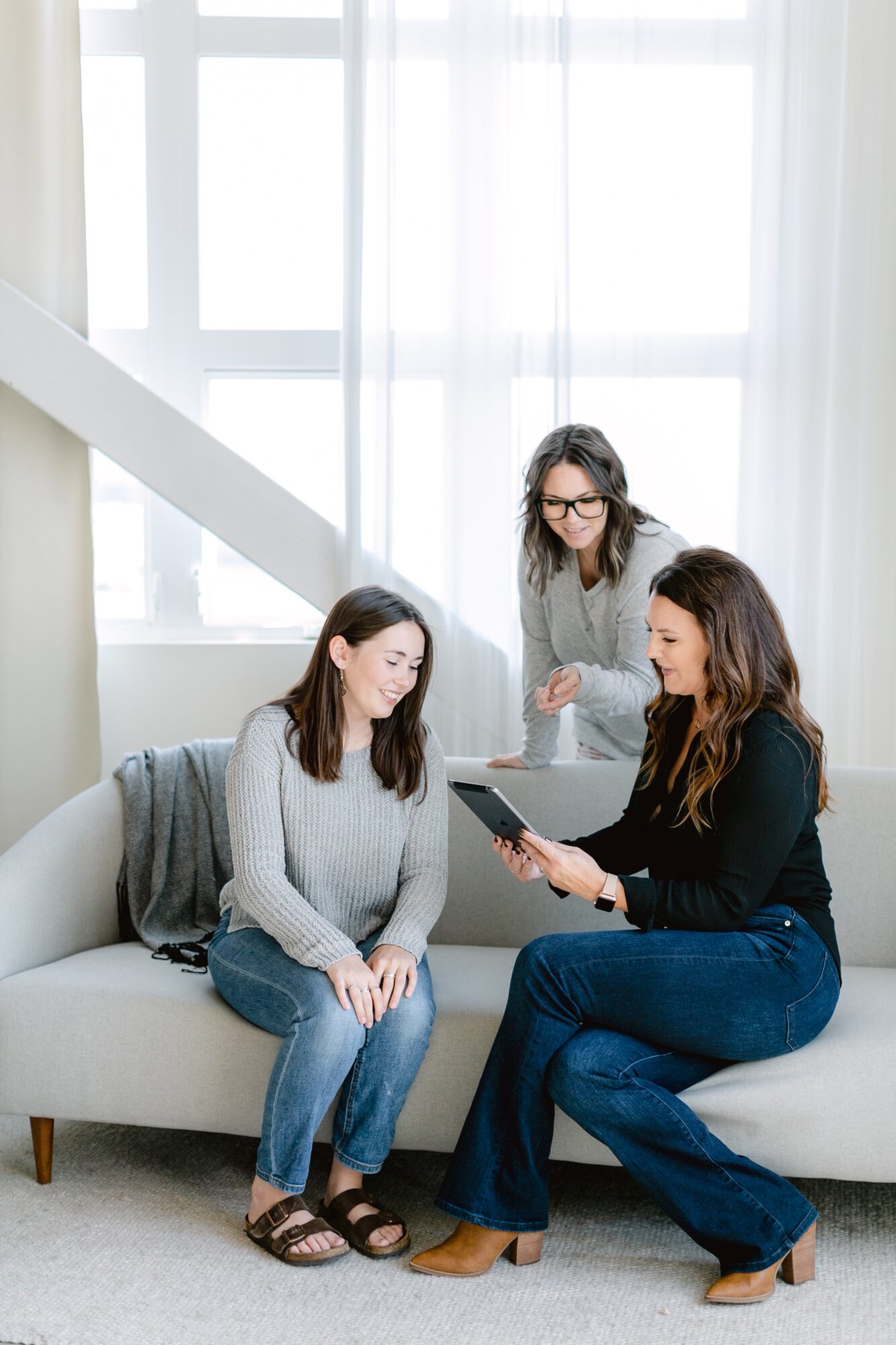 Three women sitting and standing in a bright room with large windows, one showing a tablet to the others.