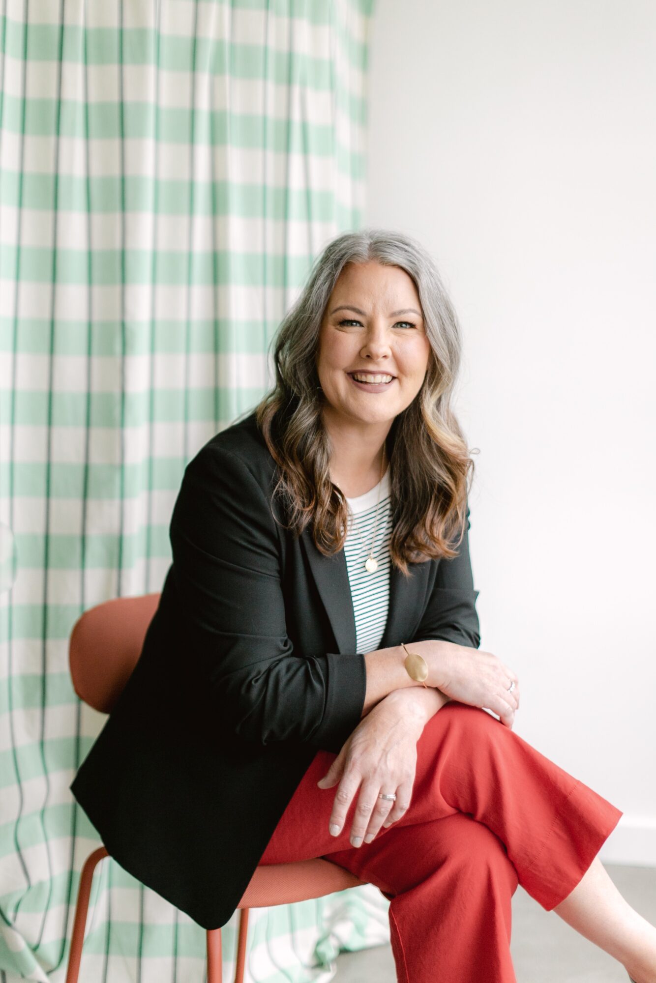Smiling woman with wavy hair, sitting on a chair, wearing a black blazer and red pants, in a room with green and white checkered curtains.