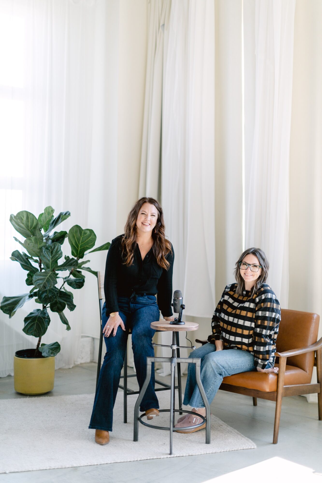 Two women in a room with white curtains, one standing and one sitting on a chair, with a large potted plant nearby.