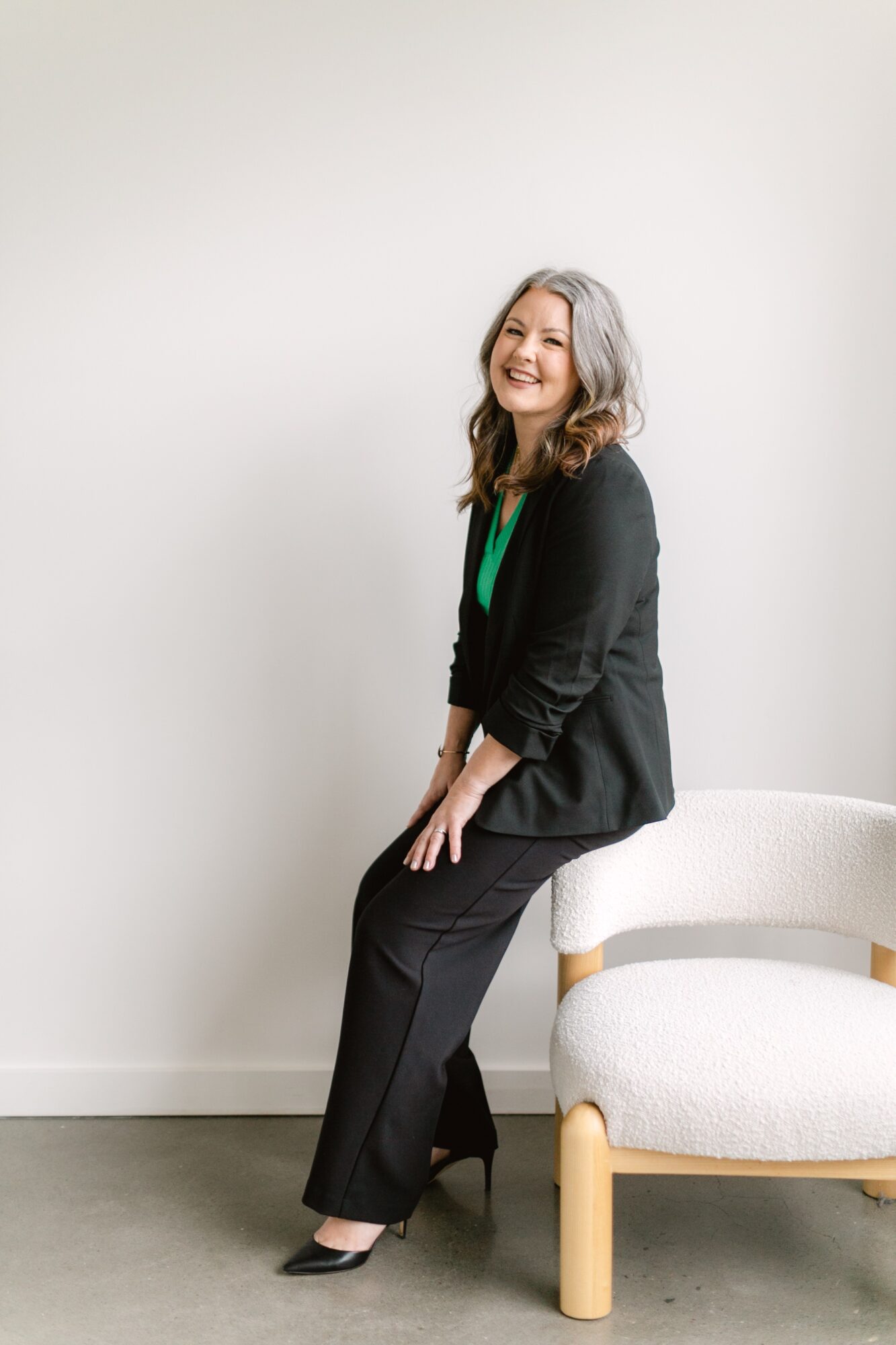 Smiling woman with gray hair standing next to a white chair, wearing a black blazer and pants, against a plain white wall.
