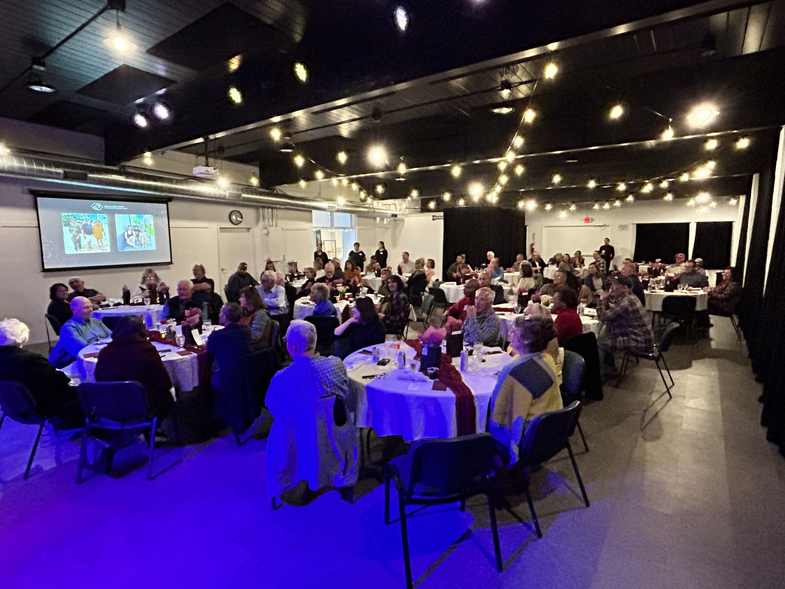 Large banquet hall filled with people seated at round tables, with a presentation screen on the wall, ceiling lights, and a dark ceiling.