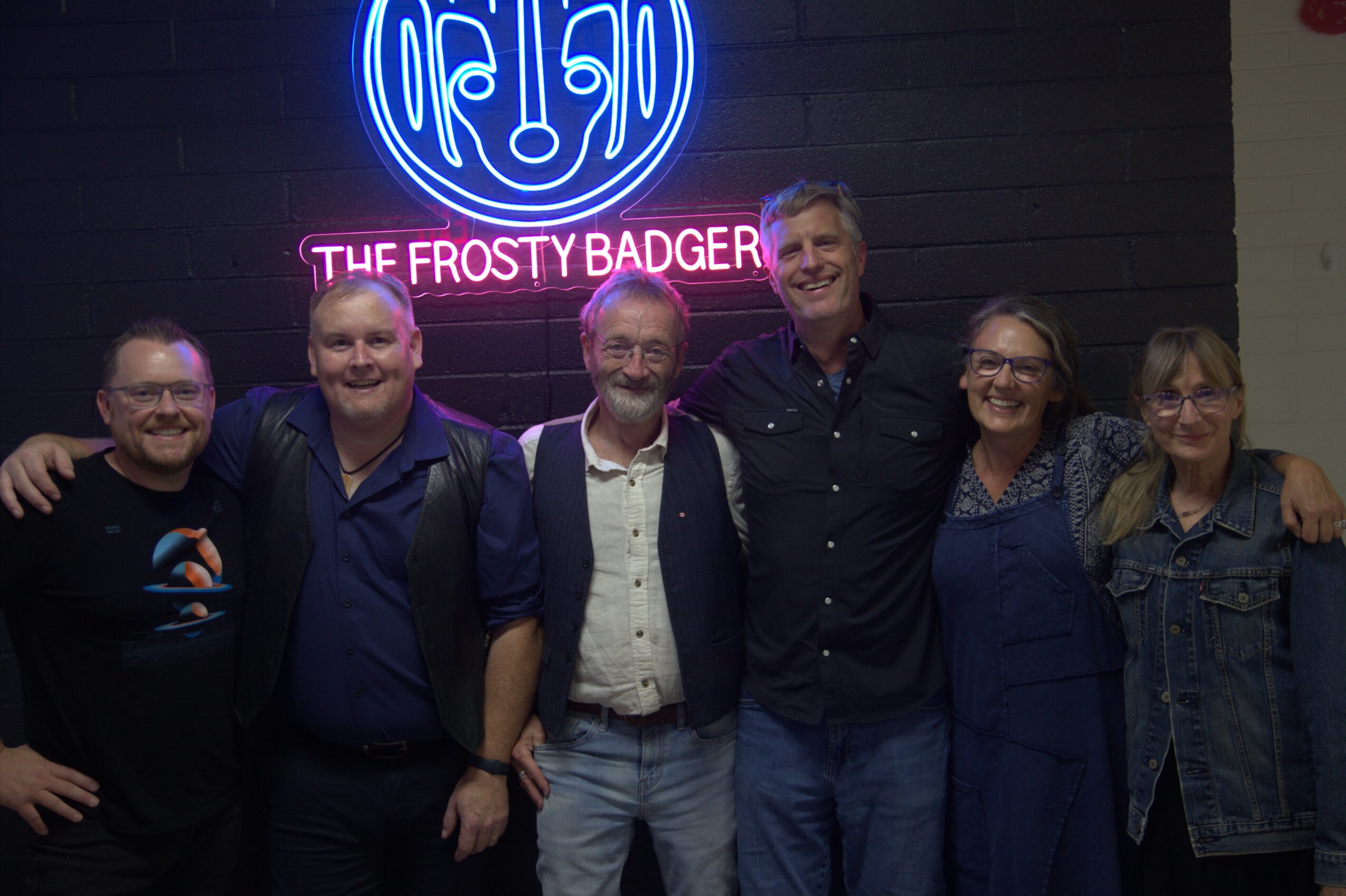 Group of six people standing together indoors, smiling, with a neon sign reading 'The Frosty Badger' behind them.