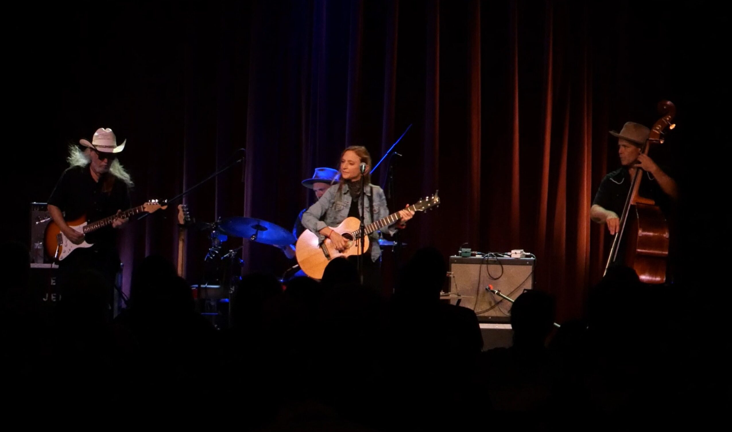 Three musicians perform on stage with guitars and a double bass, illuminated by stage lights, against a dark background.