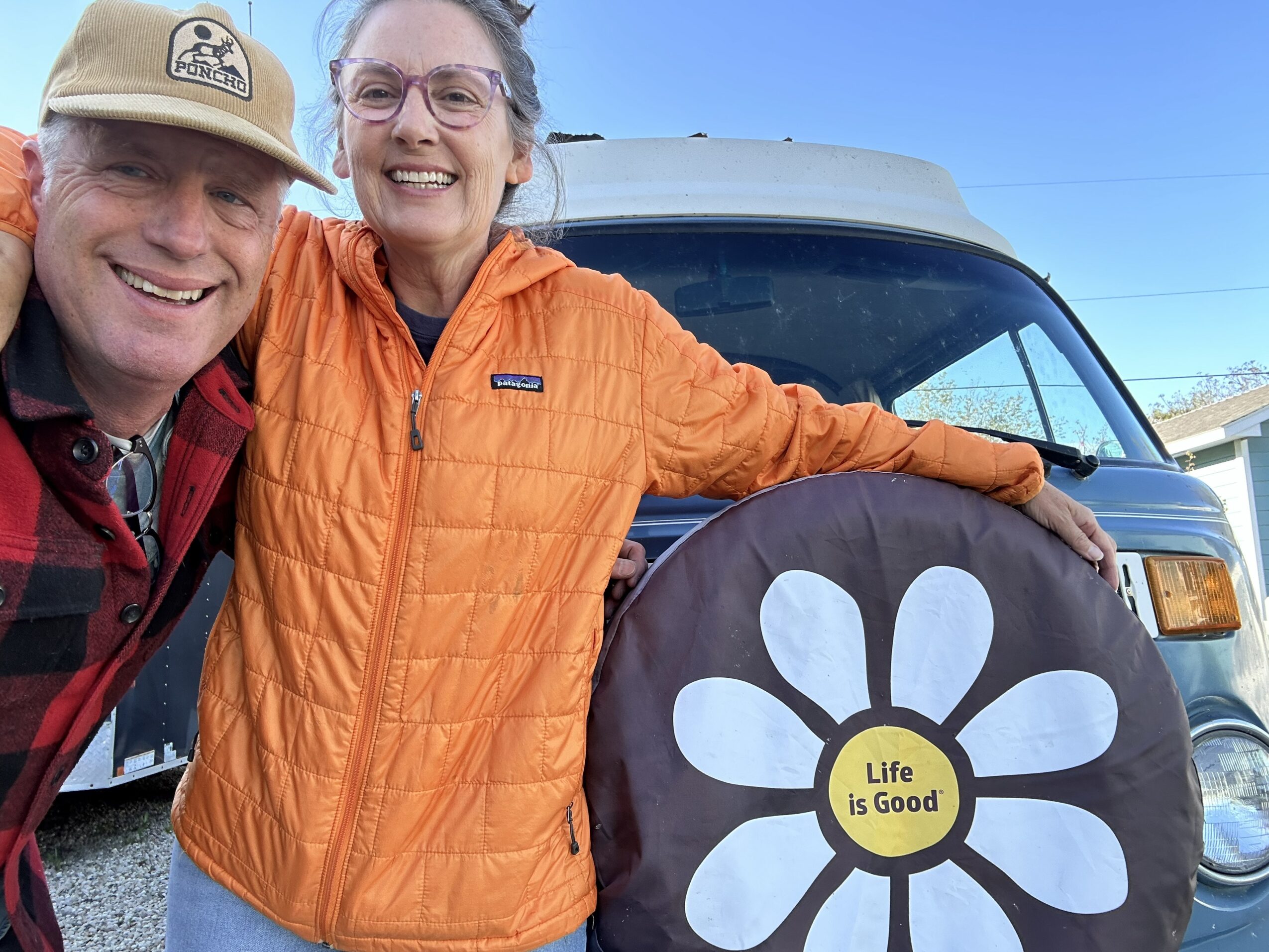 Two smiling people, a man and a woman, stand outdoors near a vehicle, with the woman holding a bag with a flower design and the text 