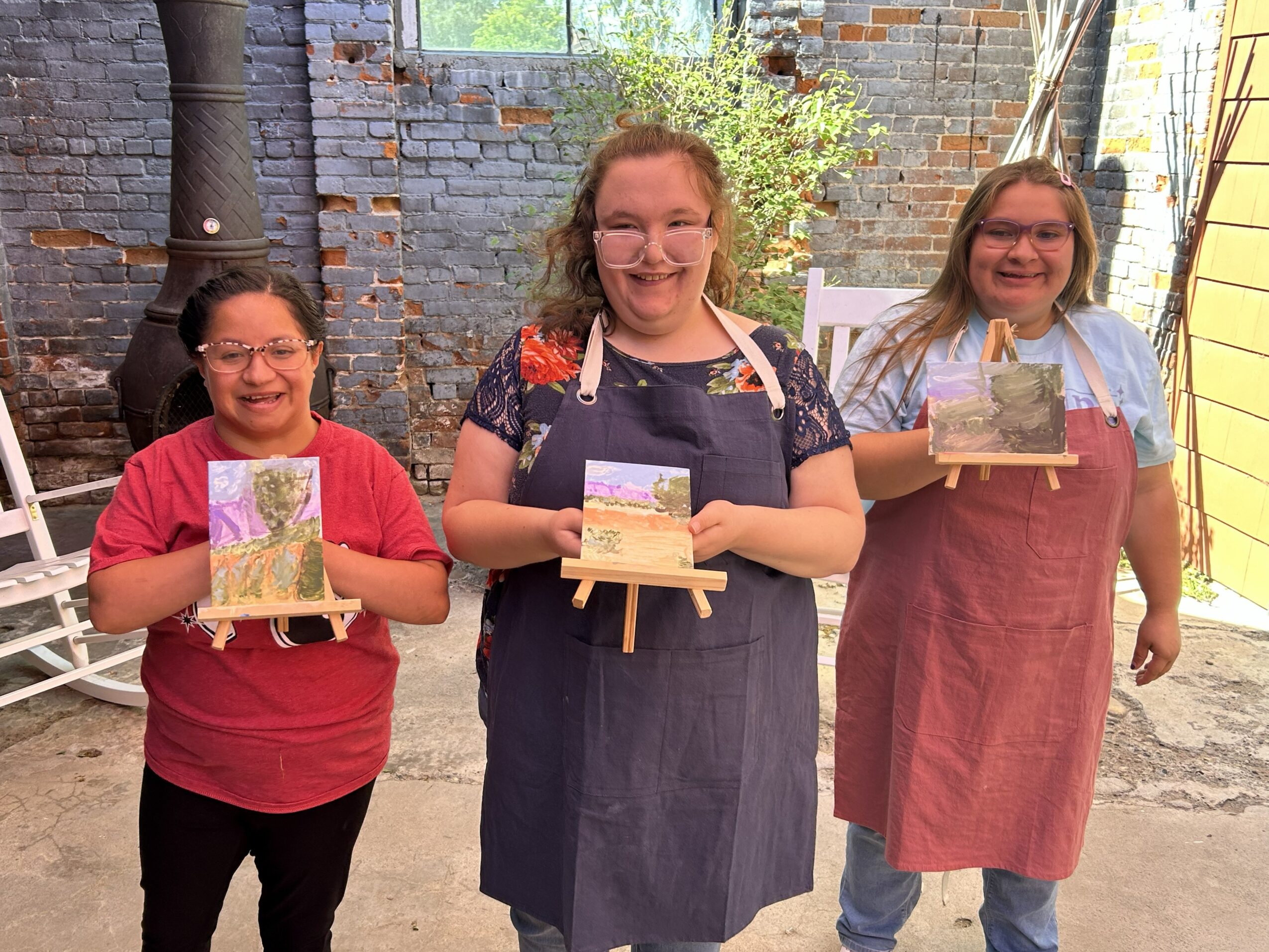Three smiling women holding small paintings in an outdoor brick setting.