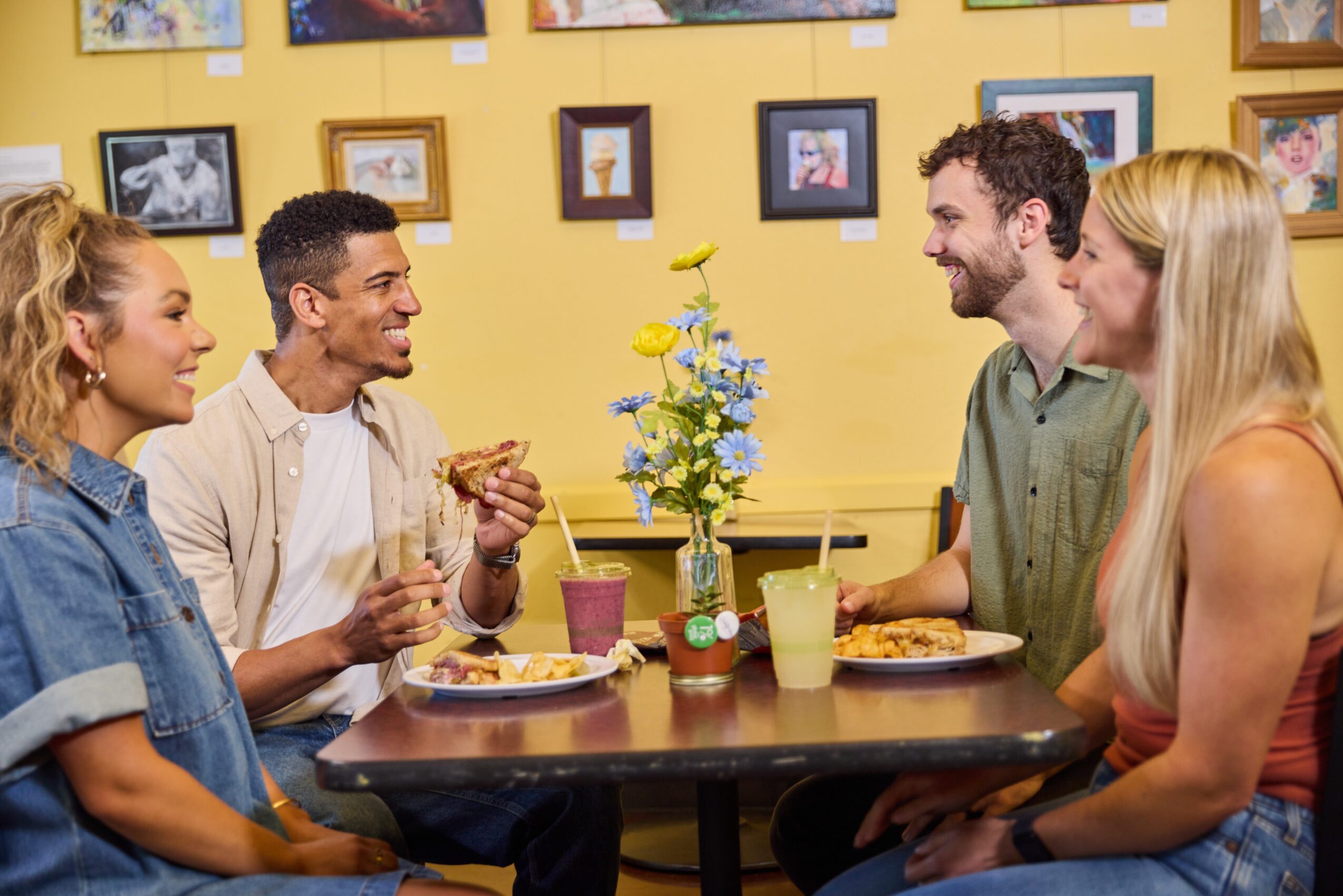 Four people sit around a table with flowers, eating and talking, in a room with framed pictures on a yellow wall.
