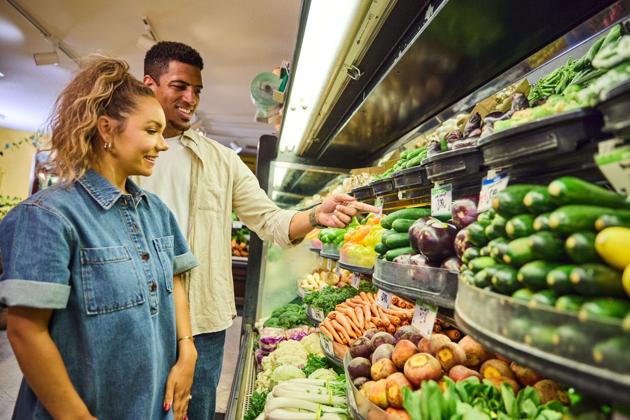 Two people shopping for vegetables in a grocery store, smiling and selecting produce from shelves.