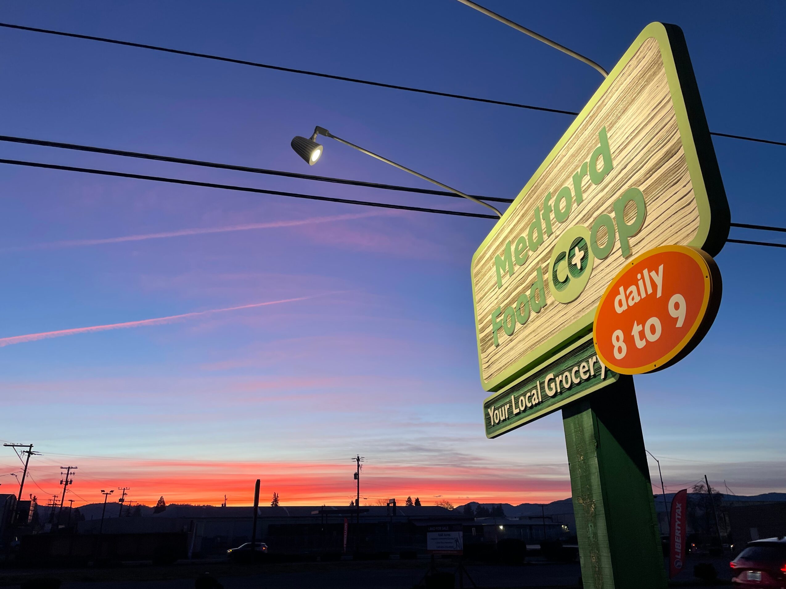 Sunset sky with a sign for a neighborhood cooperative store, streetlights, and power lines, with a silhouette of buildings in the distance.