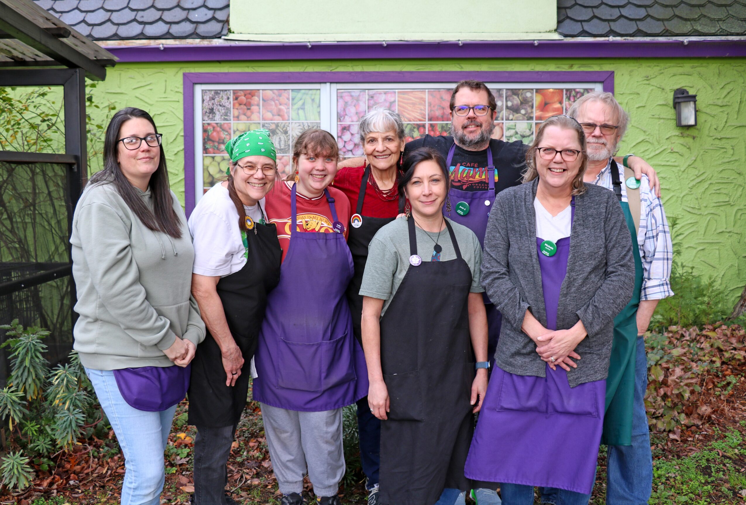 Group of nine people standing outside in front of a green building with a window, smiling, some wearing aprons and glasses.