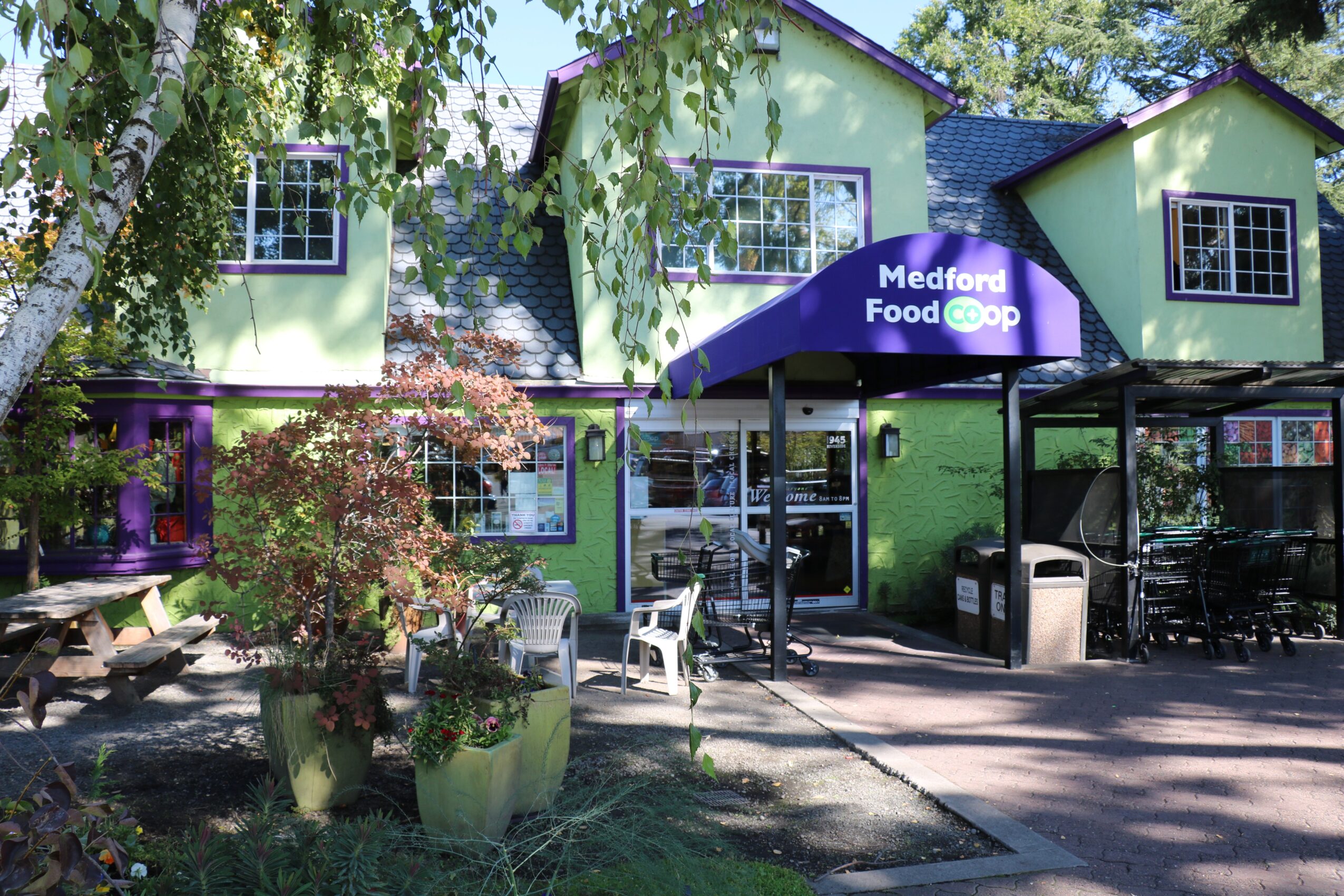 Green building with a sign reading 'Medford Food Co-op,' outdoor seating, trees, and plants in front.