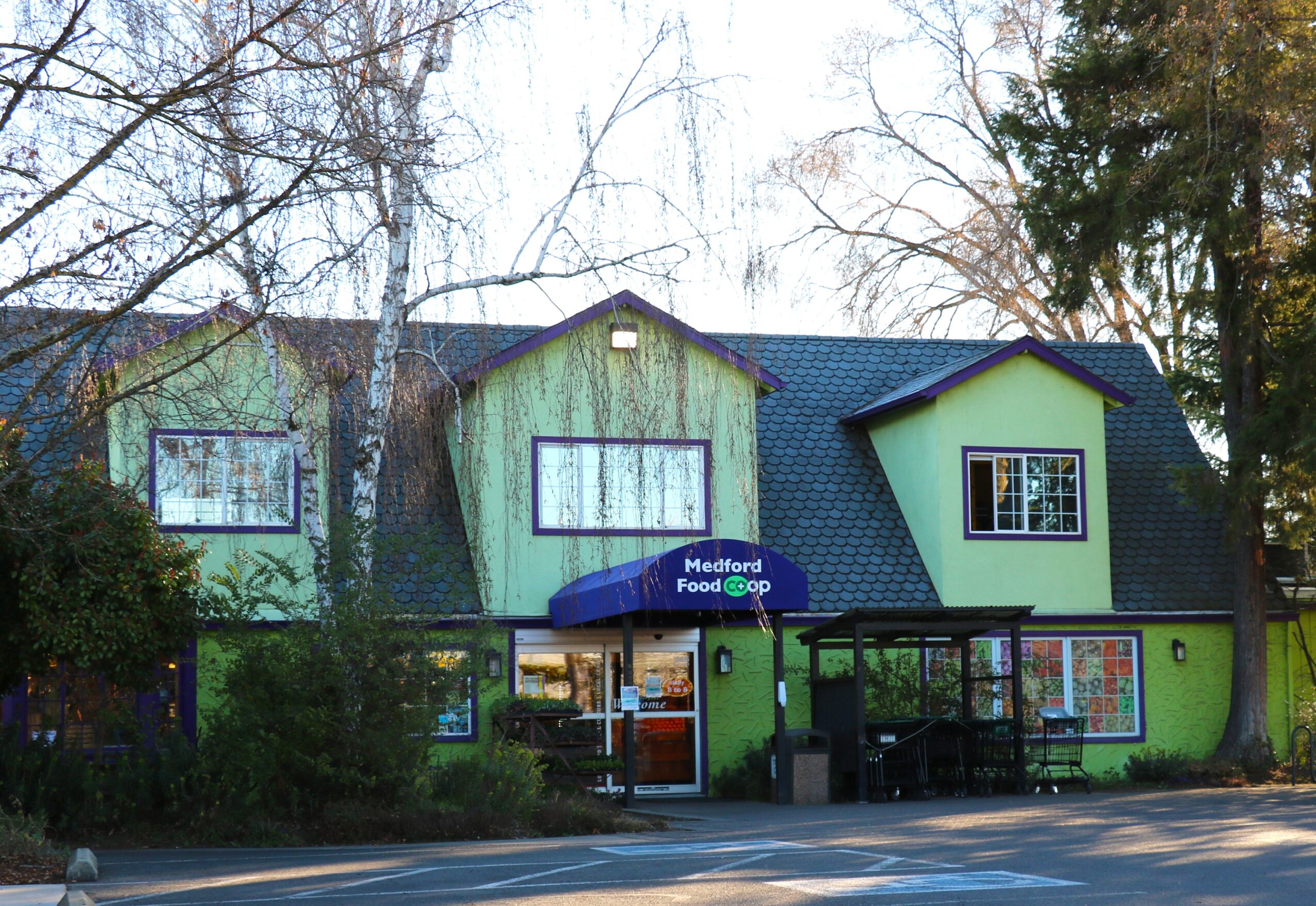 Green building with multiple windows and a blue sign reading 'Medford Food Co-op' in front, surrounded by trees.