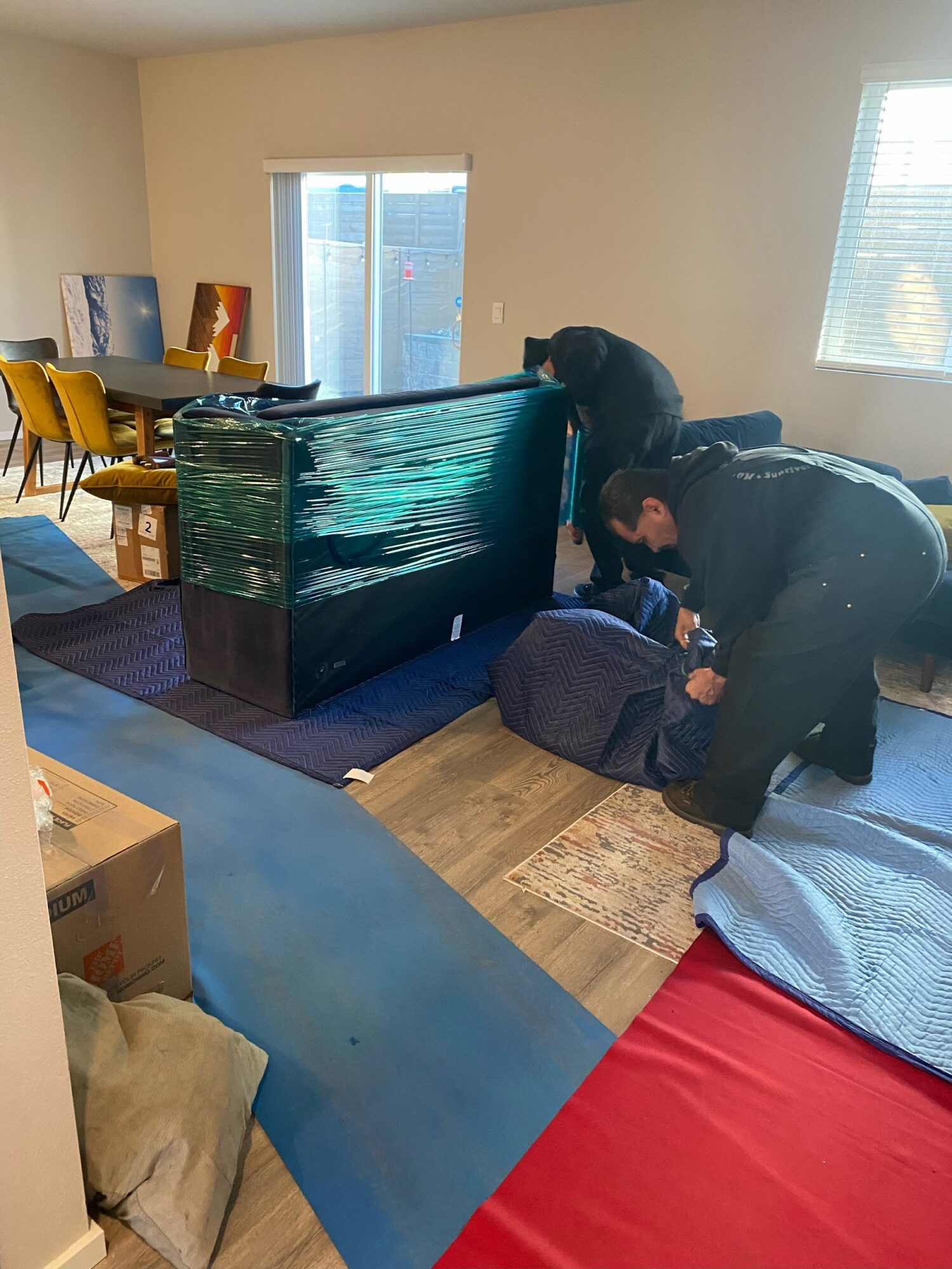 Two people unpack a large flat-screen TV from plastic wrap in a living room, with a dining area and windows in the background.