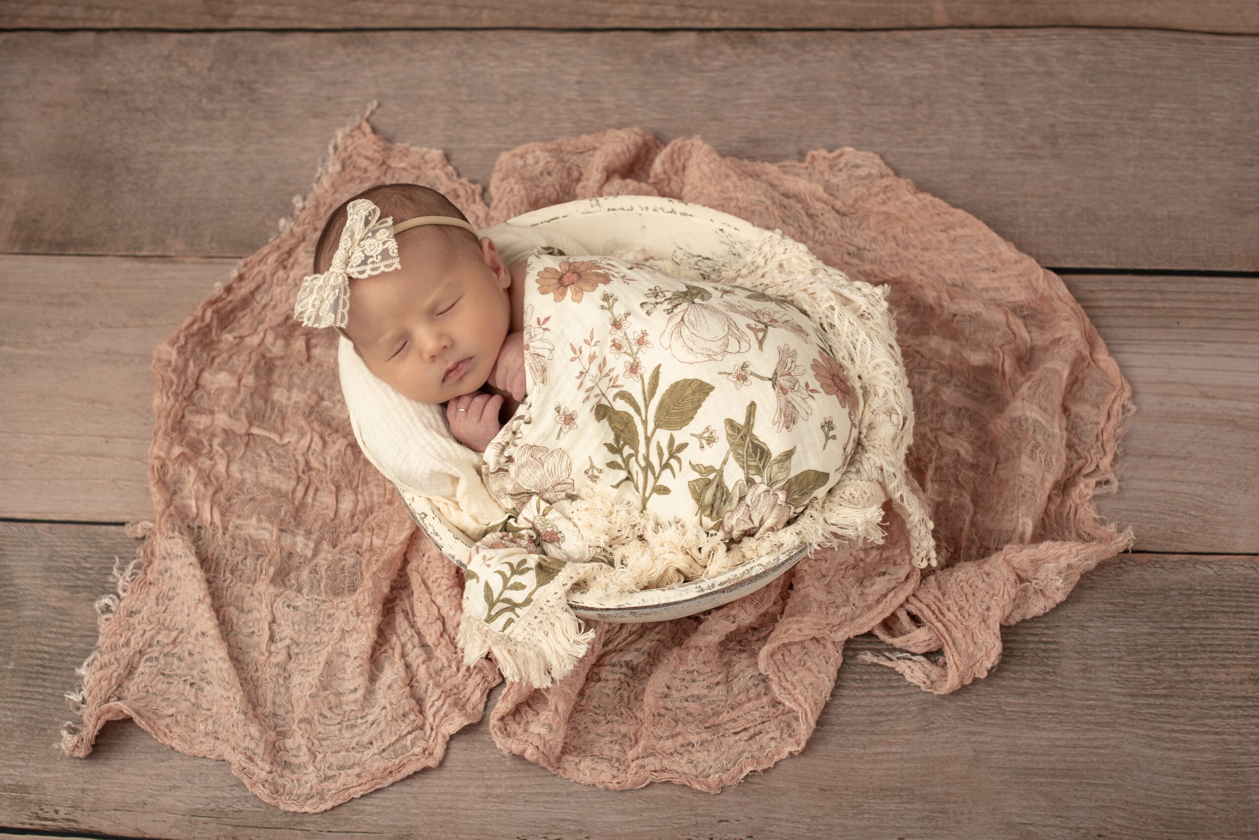 Newborn baby sleeping on a blanket, wrapped in a floral cloth, with a headband, on wooden floor, 20 words