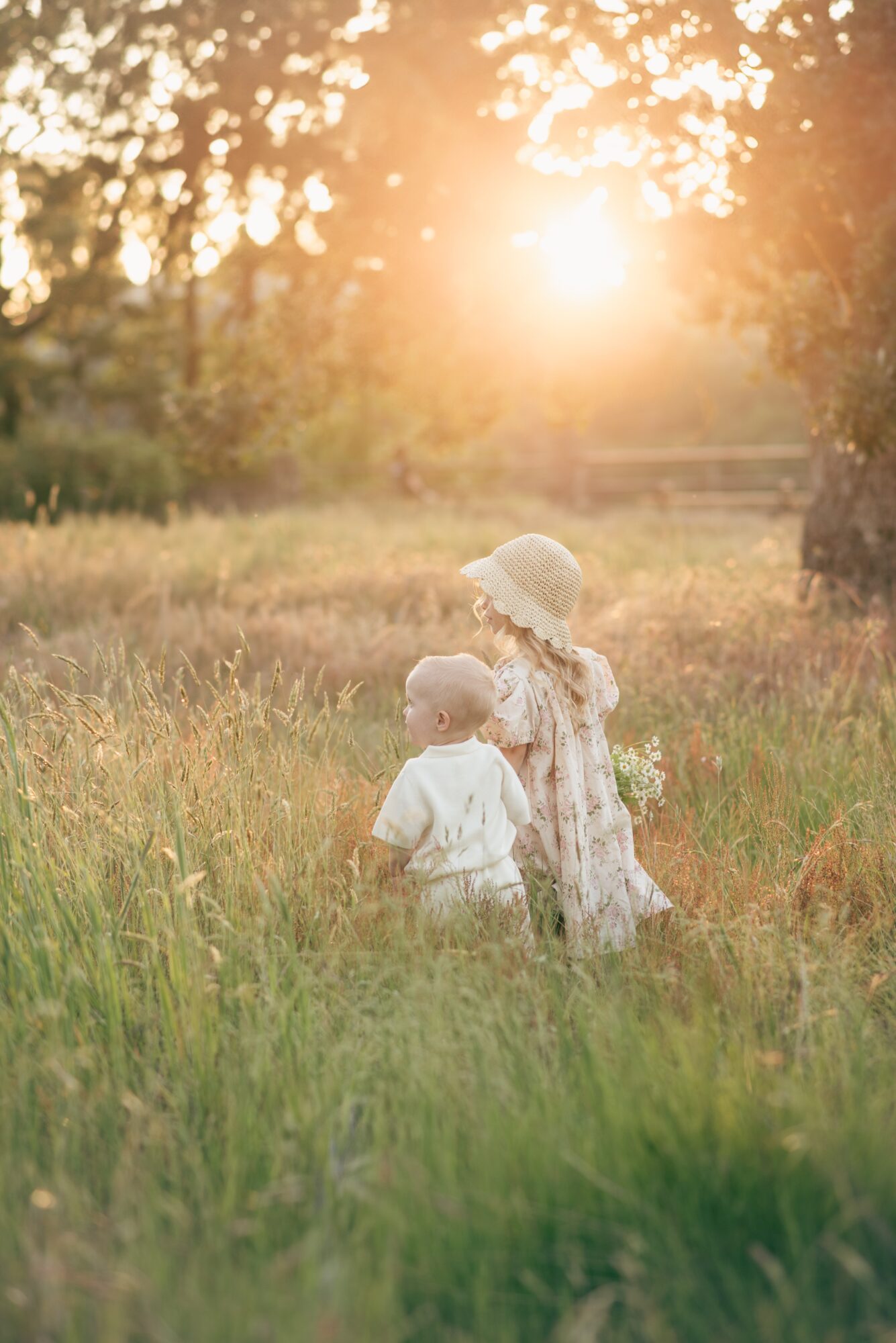 Two children in a grassy field with trees and sunlight in the background, one wearing a hat and the other facing away.