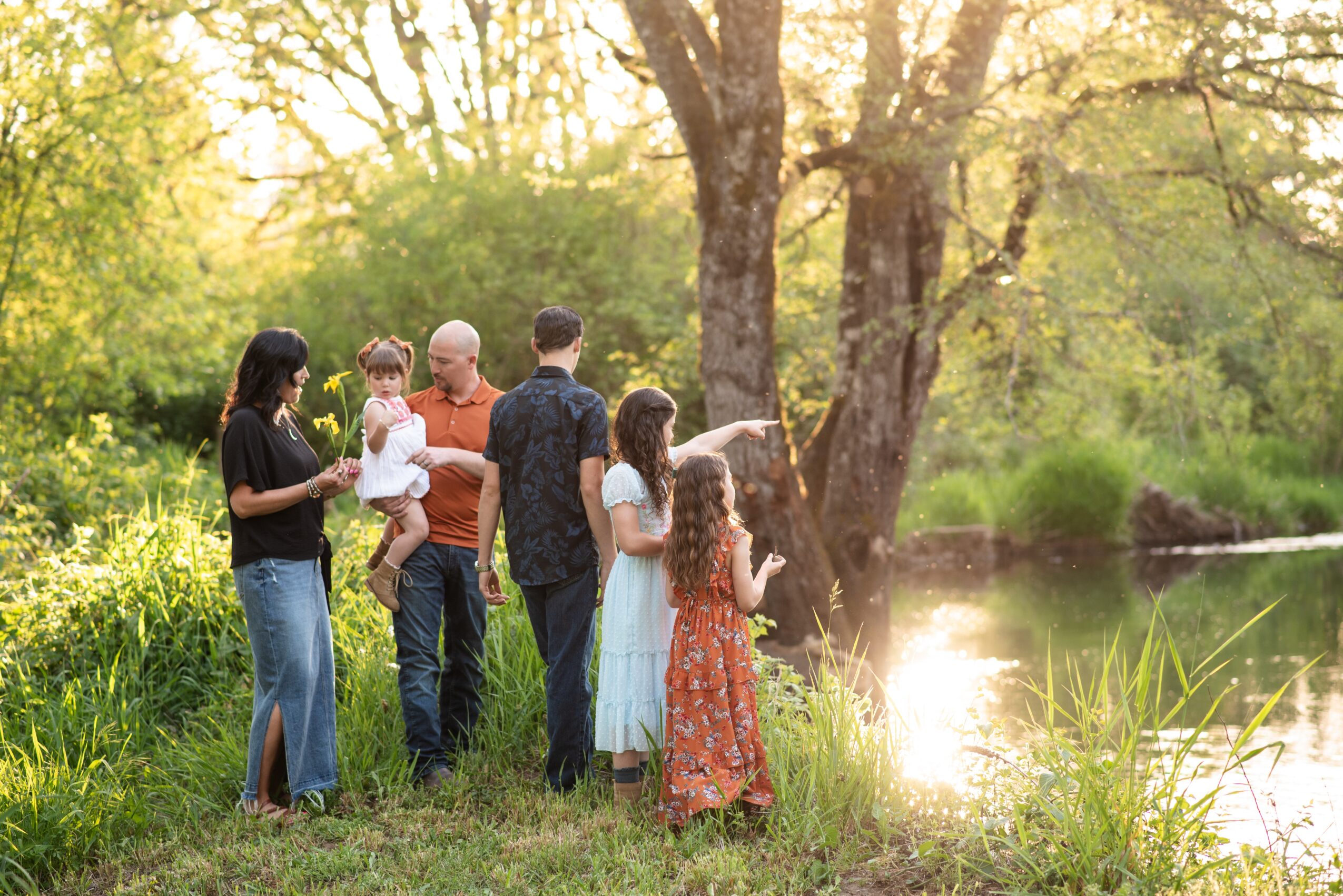 Group of people standing by a riverbank in a park with trees and greenery, enjoying a sunny day.