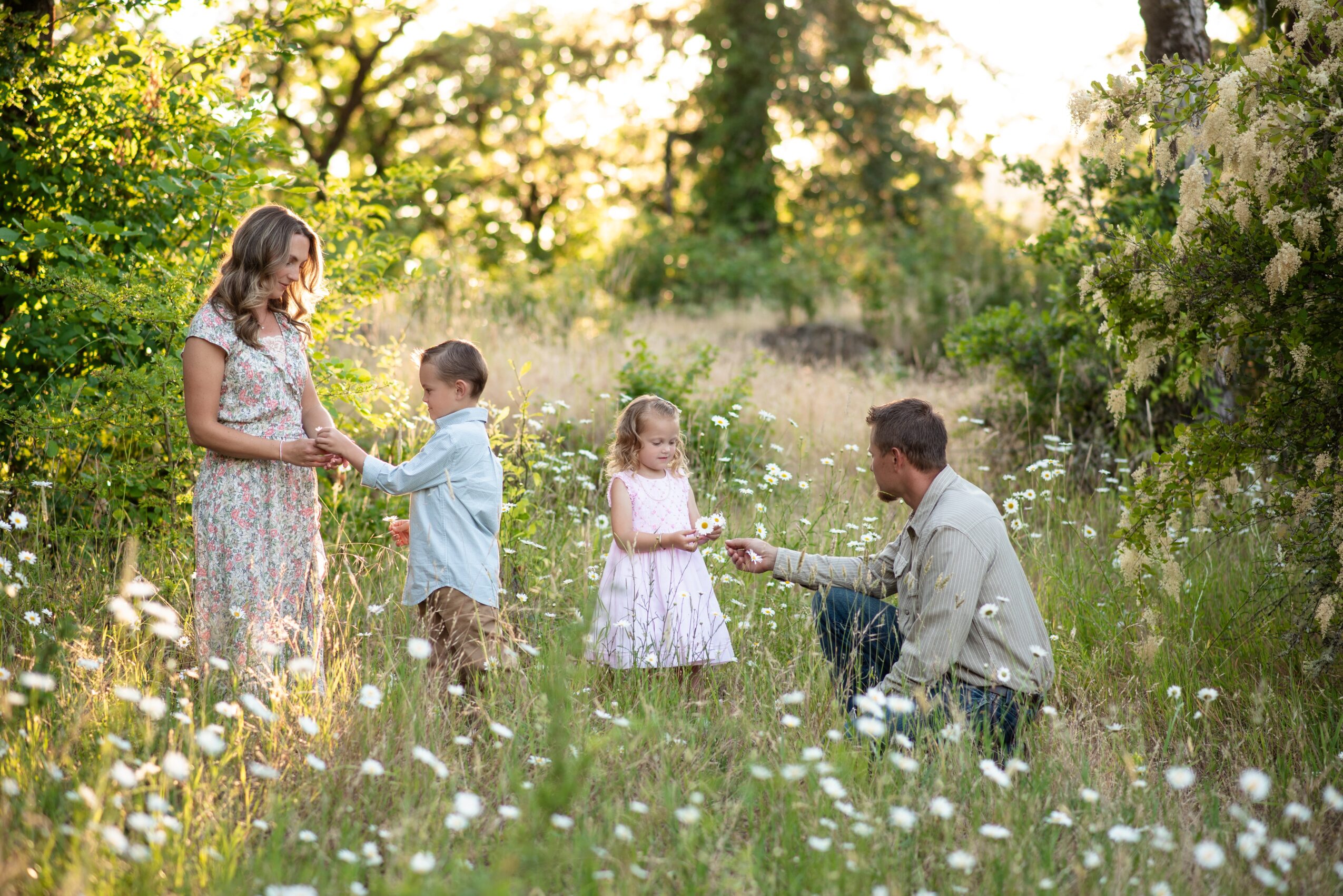 A woman and three children in a grassy field with trees, playing with water spray, surrounded by flowers and greenery.