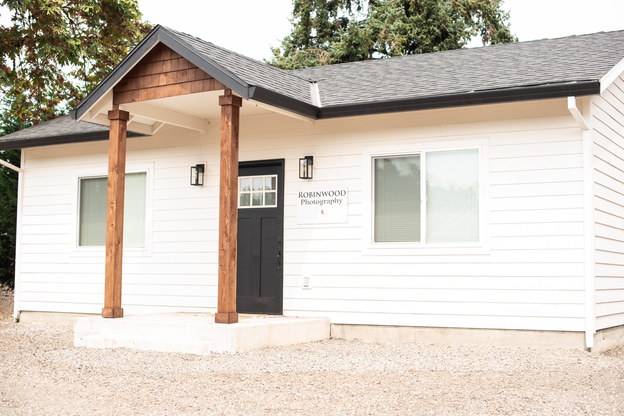 White house with black door, two windows, and wooden porch columns, surrounded by gravel and trees in background.