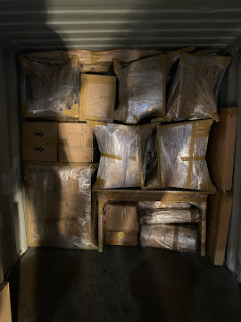 Stacked boxes wrapped in plastic inside a storage unit, with some boxes on top of others and a dark floor.