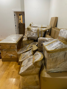 Room filled with wrapped furniture and boxes, some stacked against a white wall, hardwood floor visible.