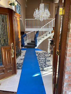 View of a hallway with a blue carpet runner, chandelier, staircase, and wooden furniture, seen from an open door.