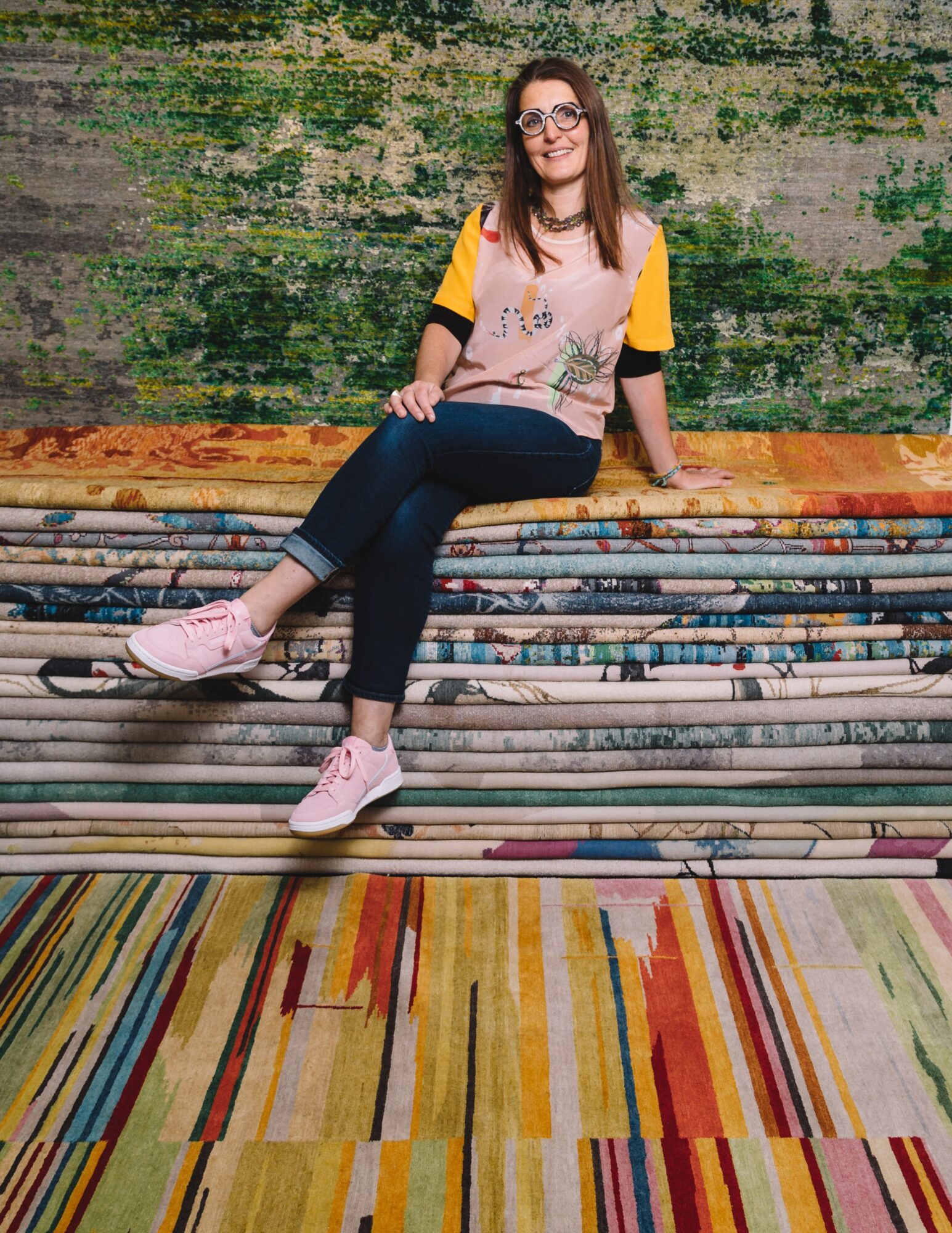 Woman sitting on colorful rolled carpets, smiling, wearing glasses, a yellow and pink shirt, dark pants, and pink shoes, in front of a green textured wall.